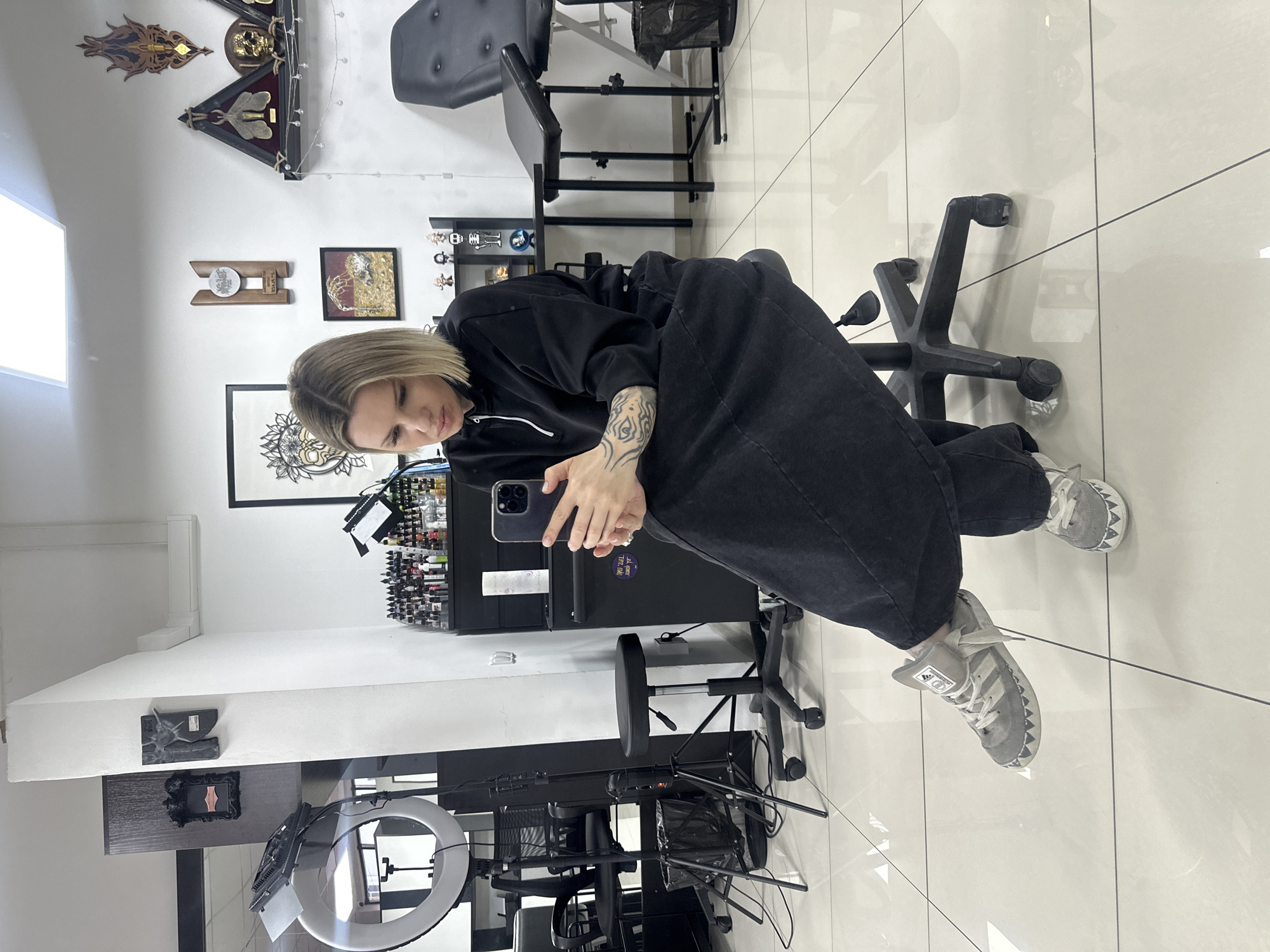 The scene is an interior view of what appears to be a tattoo studio located in Rostov-on-Don, Russia. The room features white walls and a light-colored tiled floor with dark grout lines. Overhead fluorescent panel lighting illuminates the space.
A blonde adult female is seated centrally on a black five-wheeled office stool, facing slightly left and looking down intently at a dark-colored smartphone held in both hands. She is wearing a black long-sleeved hooded top, dark loose-fitting trousers, and grey Adidas sneakers with a distinctive white serrated pattern along the sole edge. Tattoos are visible on her right forearm. Her left leg is crossed over her right at the ankle.
To the left of the seated individual, a large circular ring light and a square LED panel light are positioned on stands. A black office chair and a black waste bin are also present. A tall, dark cabinet or partition partially separates the space.
The background displays a black shelving unit or cabinet loaded with numerous small bottles of tattoo ink in various colors, alongside other supplies. The walls are adorned with framed artworks, including a large intricate drawing, a wooden award plaque, and a smaller framed piece. Further right, a triangular display case containing a mounted insect specimen and a golden skull is visible, along with an ornate wooden wall carving and a string of decorative lights. A white wall-mounted air conditioning unit is positioned high on the right wall.
To the right of the seated individual, a black adjustable tattoo chair with a padded seat and back is present, accompanied by a small black adjustable side table. A black rolling cabinet with a roll of white paper towels on top is positioned near the tattoo chair.