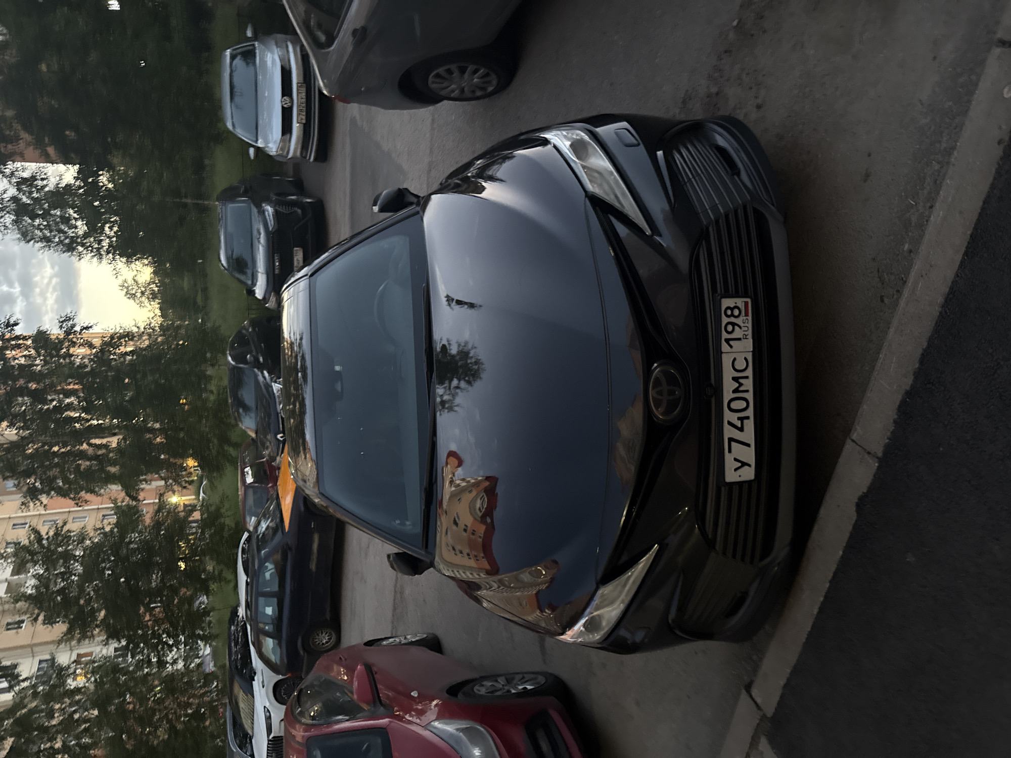 The image shows a black Toyota Camry parked in a residential courtyard in Saint Petersburg, Russia. The car is in the foreground, facing the viewer, with its headlights on. The license plate is visible and reads "Y 740MC 198 RUS". In the background, several other cars are parked, including a red Kia Rio, a dark blue station wagon with a yellow hood, a silver Volkswagen Jetta, and a black SUV. Apartment buildings and trees are visible behind the cars. The sky is overcast, suggesting it is either early morning or late evening. The ground is asphalt and concrete. There are no people or animals visible in the image.