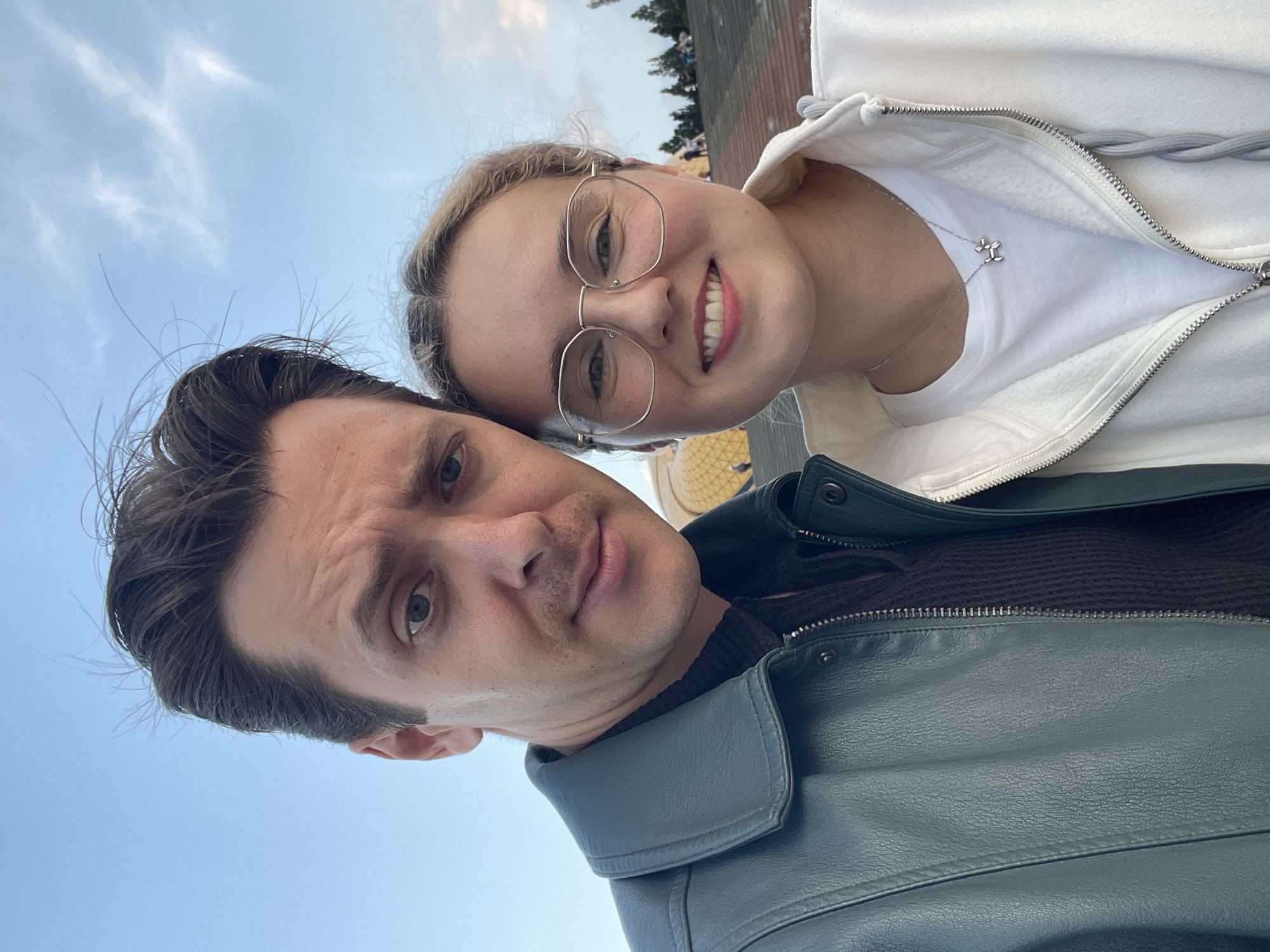 Two people pose for a selfie against a blue sky. A man with dark hair and a green leather jacket is in the foreground. Behind him, a woman with glasses and a white zip-up jacket smiles. The background reveals a glimpse of the city of Da Lat, Vietnam, including a notable architectural structure. The individuals are positioned close together, likely interacting warmly.