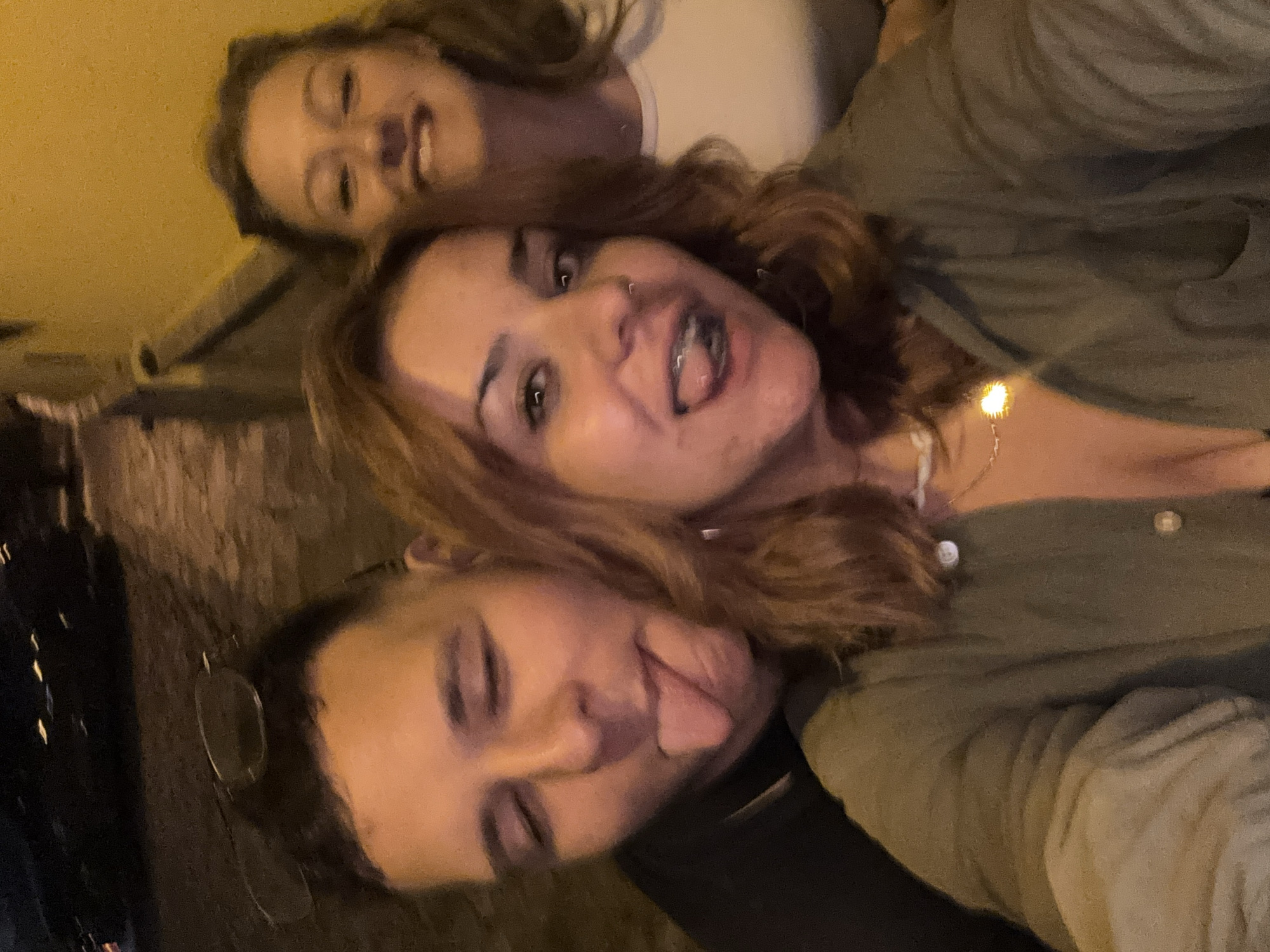 This is a close-up, selfie-style photograph featuring three young women in what appears to be an outdoor setting at night. The lighting is warm and somewhat dim, suggesting artificial light sources like streetlights or a string of fairy lights, which are visible as small glowing orbs in the middle right of the frame. The women are positioned in a cluster, with their faces filling most of the frame.
The woman in the foreground on the left, wearing glasses, has her head tilted back, and her mouth is slightly open, showing teeth. She is wearing a dark garment. Above her and slightly to the right, a woman with auburn hair is looking towards the camera with her tongue sticking out, her mouth open in a playful expression. She is wearing a light-colored necklace and a green top. Partially visible behind and above her is a third woman, whose face is less clear but also appears to be looking at the camera with a smiling expression. She has blonde hair and is wearing a white top.
The overall mood of the photograph is casual and convivial, with the women appearing to be enjoying themselves. The background is dark and indistinct, with some texture that could be a wall or natural ground. There are no visible text elements in the image. The scene is consistent with a social gathering or outing in Plovdiv, Bulgaria, during the evening.