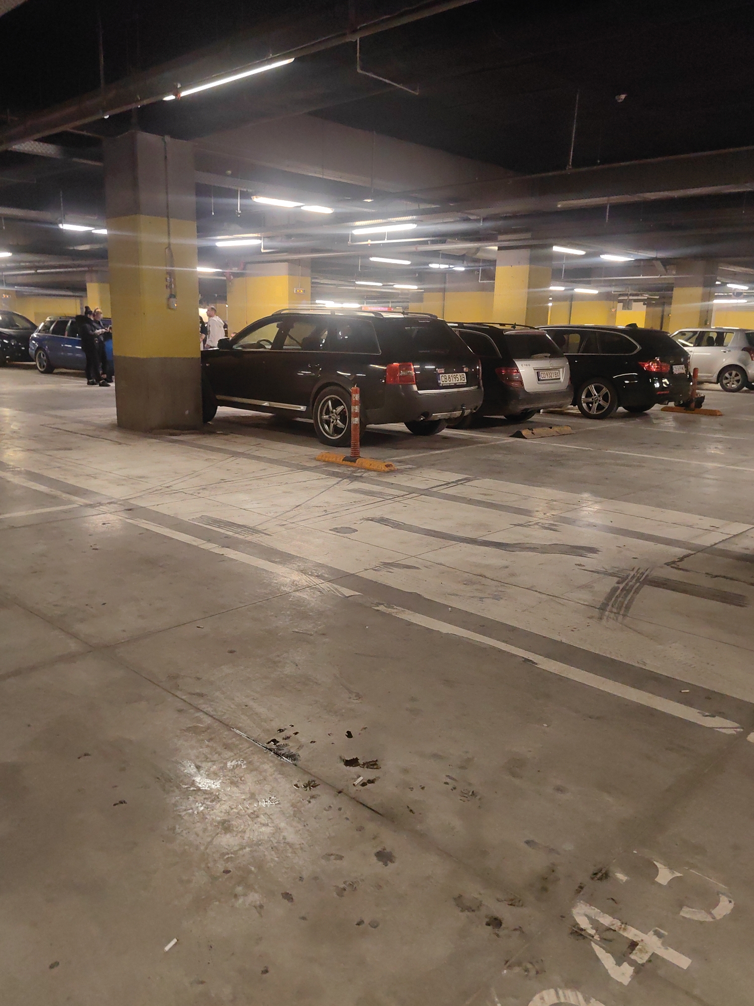 The image depicts an indoor parking garage, likely located in Sofia, Bulgaria based on the visible license plates. Fluorescent lights illuminate the garage, casting a cool tone over the scene. A row of parked cars is visible, including a dark-colored Audi with license plate "CB 8195 XB", a silver Mercedes-Benz with license plate "C09321BX", and several other vehicles. Some individuals are standing near a large, yellow support column, possibly interacting with one another. The ground is concrete, showing signs of wear and some dark spots. Parking lines are painted on the floor.