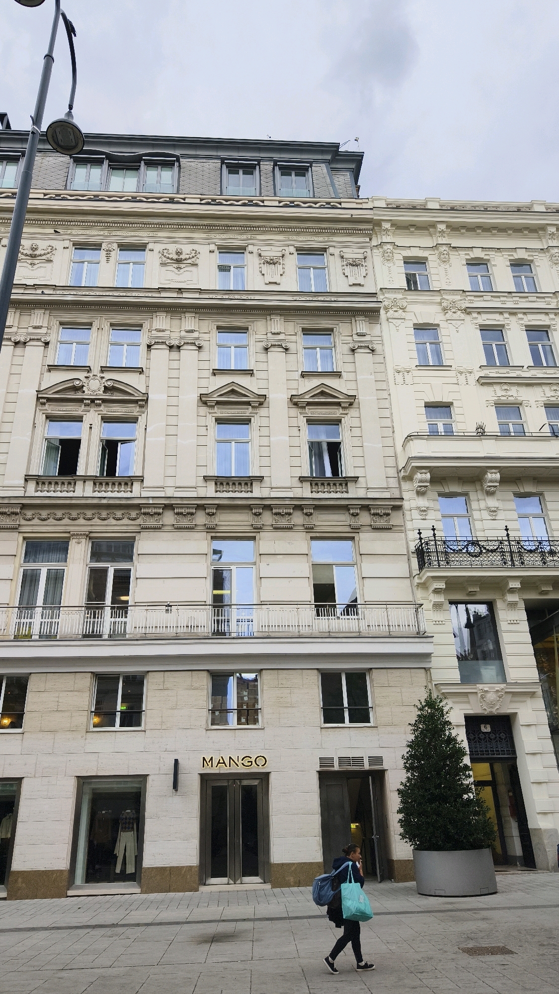 A low-angle medium shot shows the facade of two adjoining buildings on a city street. The building on the left has a mansard roof with dark gray, diamond-shaped tiles and several dormer windows. The building on the right has a lighter-colored, ornate facade with classical architectural details, including balconies with black wrought-iron railings. The ground floor of the left building features a "MANGO" store with large glass windows and double doors. A woman walks across the sidewalk in front of the store, carrying two shopping bags, one light blue and the other dark blue. A large potted evergreen tree stands to the right of the store. A street lamp is visible in the upper left corner of the frame. The scene appears to be set in Vienna, Austria, due to the architectural style and the presence of the "MANGO" store, which is a popular international fashion retailer often found in European cities. The sky is overcast.
