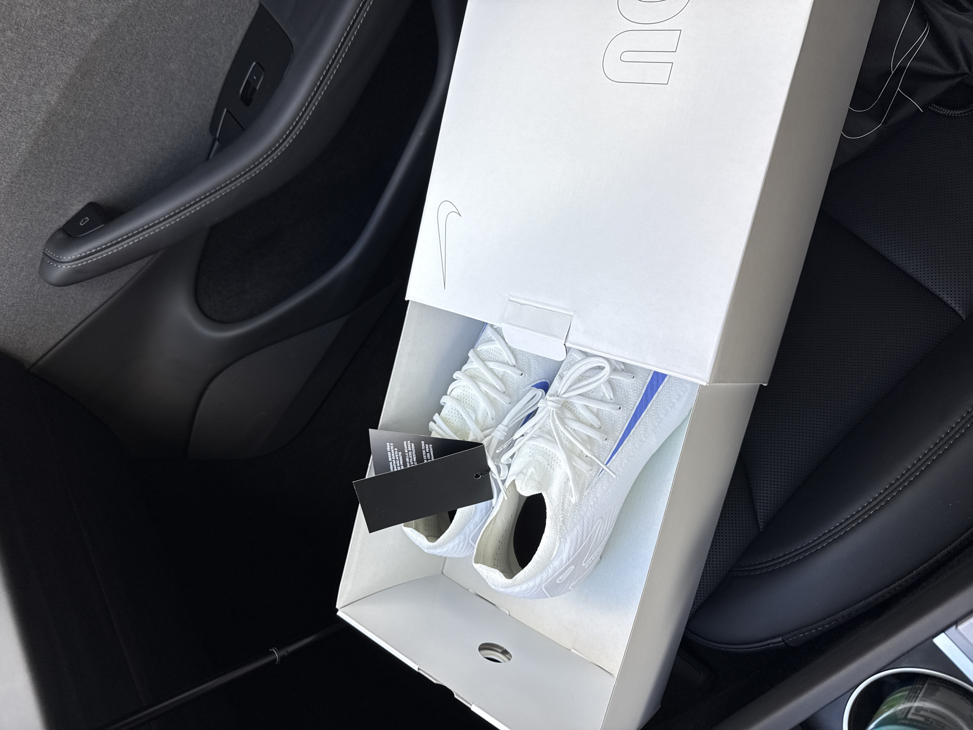 A pair of new white athletic shoes with blue accents is presented in an open white shoebox, placed on the passenger seat or floor of a car. The shoes feature white laces and a knit-like upper. A black product tag, displaying text in multiple languages such as "GASTAR DURANTE", "AFETA O", "DELLA SCARPA", "PRESTAZIO", "ESTOS TENIS", "USO. PERD", and "MIENTO", hangs from one of the shoes. The shoebox lid shows a faint outline of a Nike swoosh logo and partially visible block letters "DU" and "U".
The setting is the interior of a modern vehicle, likely during daytime, given the bright illumination. The car features black leather seating, possibly perforated, and a dark gray door panel with a distinctive black handle and white stitching. A small black button is visible on the door handle. The overall interior design suggests a sleek, contemporary car, possibly a Tesla given the door handle's specific design. The shoes appear to be newly acquired or in transit. This scene takes place in Los Angeles, United States of America.