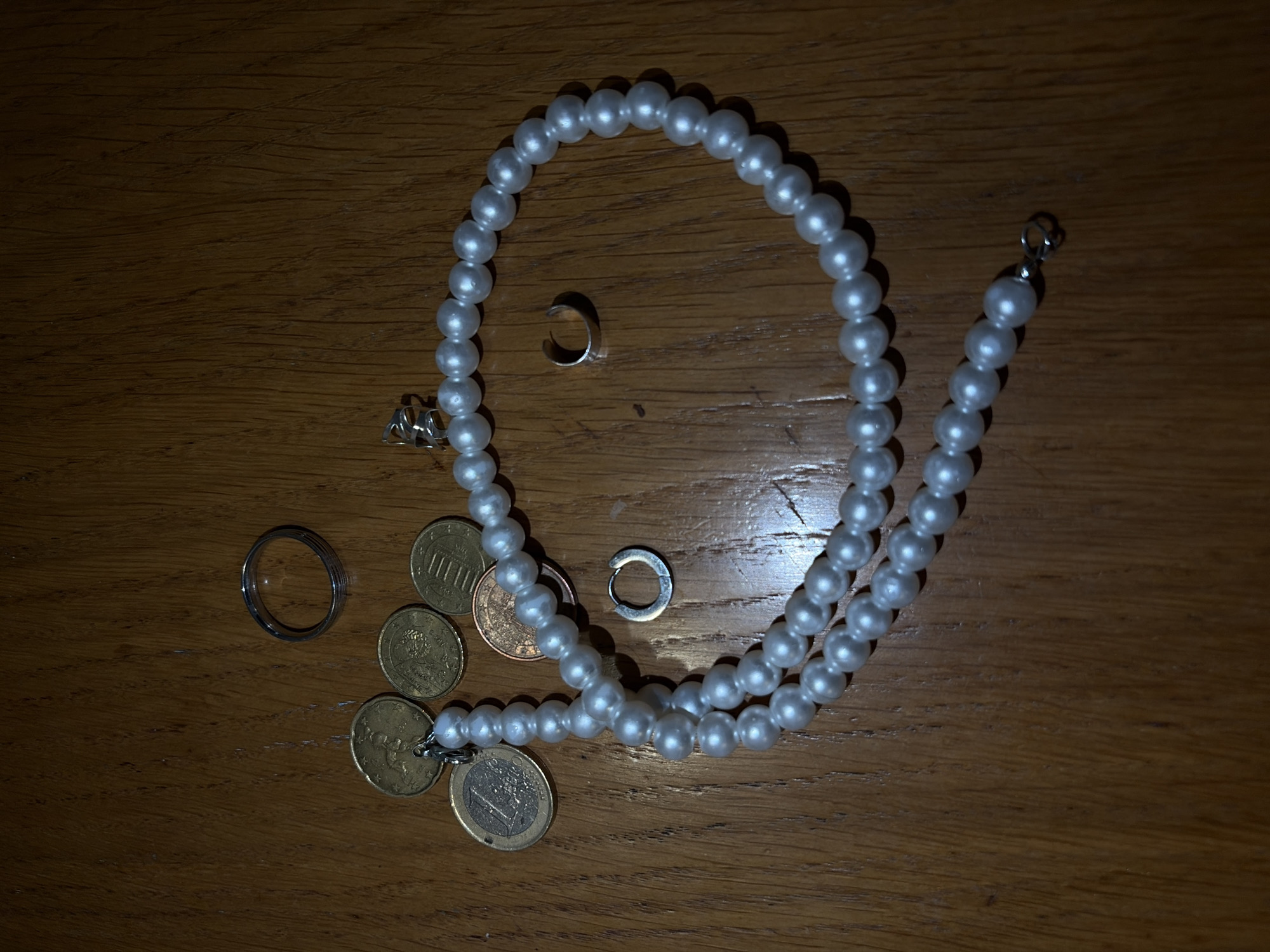 On a dark wooden surface, likely a table, lies an assortment of objects. A double-stranded pearl necklace curves across the center. Scattered around the necklace are several euro coins of varying denominations, along with a few silver rings. The lighting is dim, casting shadows and highlighting the reflective surfaces of the pearls and metal objects. No people are visible in the image. The location is unknown; no elements in the image suggest Budva, Montenegro.