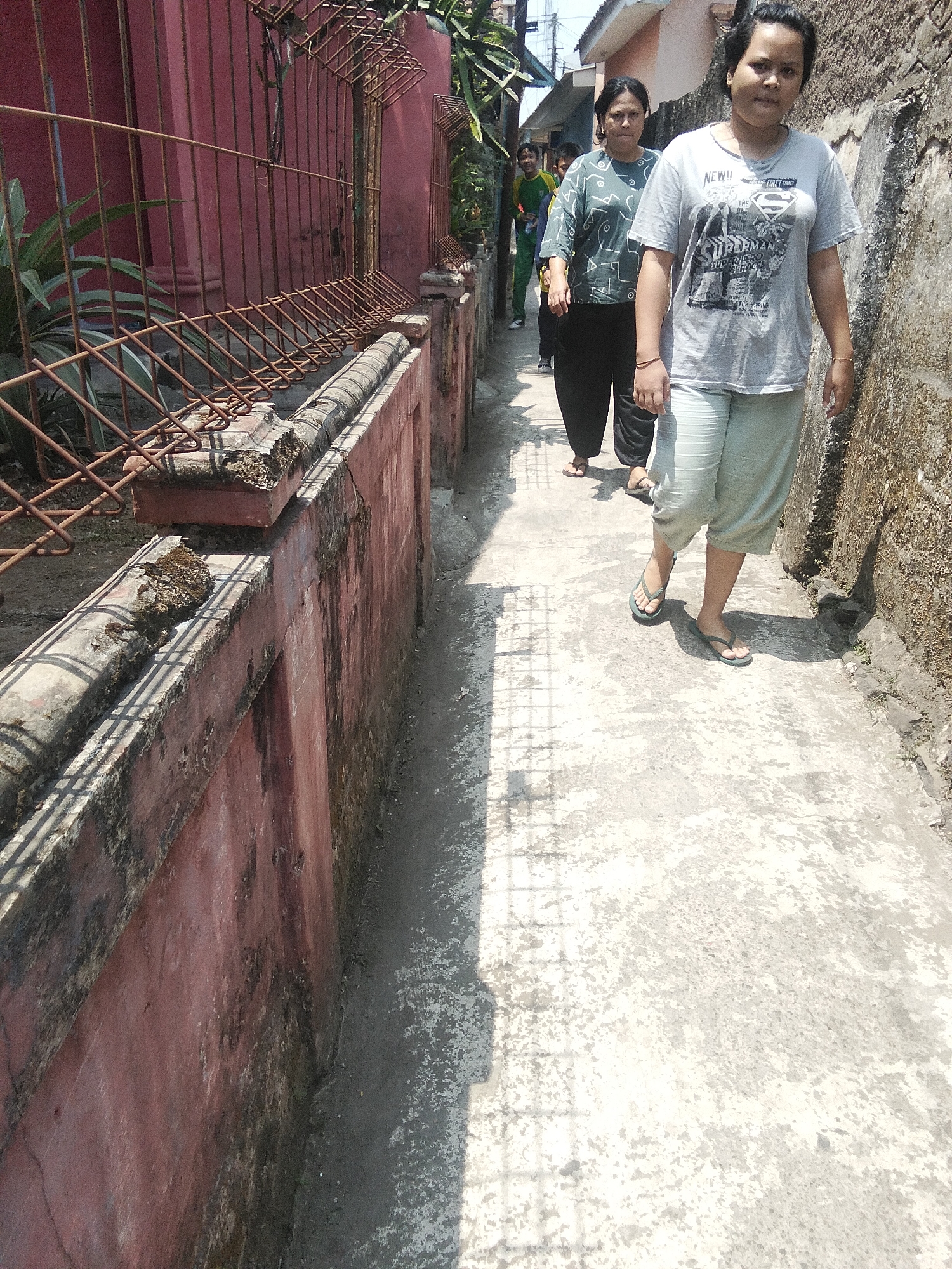 In the city of Cisaat, Indonesia, four individuals are captured walking along a narrow, sun-drenched alleyway. The alley is flanked by a weathered, reddish-pink wall on the left, topped with a corroded metal fence. On the right, a textured, light-colored wall rises, showing signs of age and possibly moss. The ground is a rough, grey concrete pathway, marked by dappled sunlight and shadows cast by unseen objects, possibly foliage or architectural elements.
The foremost person, a woman with dark hair, is wearing a grey t-shirt featuring a "Superman" graphic and light-colored capri pants. She walks with her head turned slightly towards the camera, wearing dark flip-flops. Behind her, another woman in a dark hijab and a green patterned t-shirt follows, also wearing dark trousers and flip-flops. Two other figures are visible further down the alley, one in a green and yellow shirt and dark pants, and another obscured individual. The overall atmosphere suggests a casual daytime stroll through a residential or less-trafficked urban area.