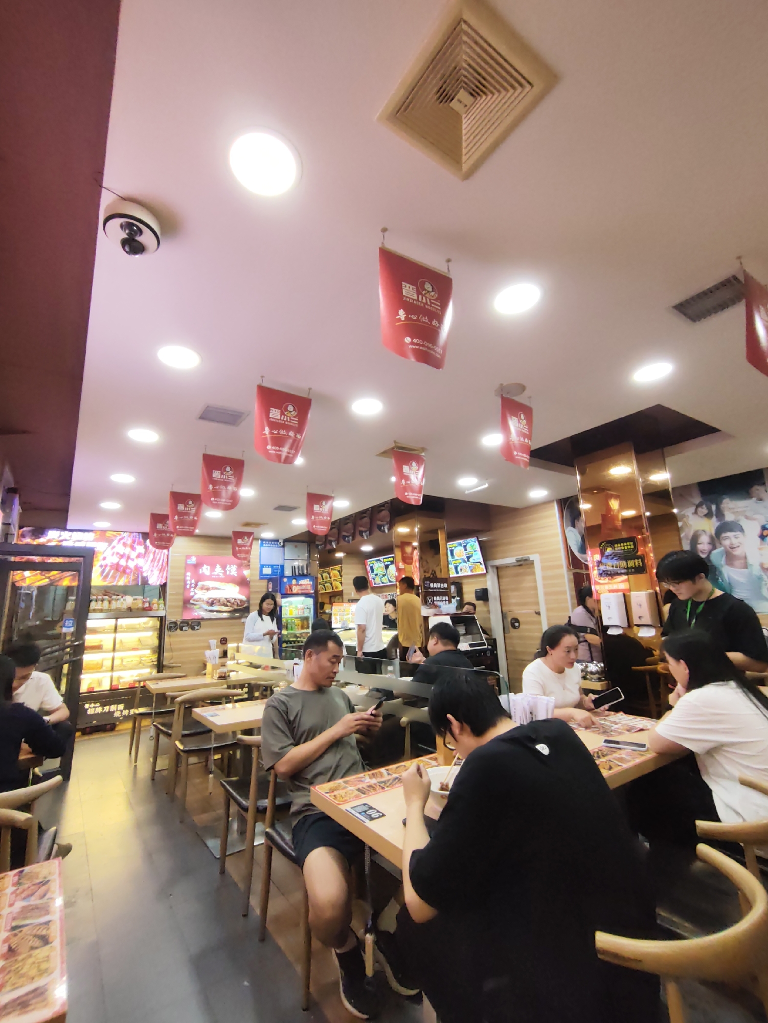 The image depicts the interior of a busy restaurant, likely a fast-food establishment, with a focus on noodles. The scene is illuminated by overhead lights, and red banners with Chinese characters hang from the ceiling, possibly advertising menu items or promotions. In the foreground, several diners are seated at tables, engaged in eating or using their phones. A man in a grey t-shirt and black shorts is seated at a table on the right, looking at his phone. In the middle ground, a man in a black t-shirt is eating from a bowl while looking down at his meal. A woman in a white shirt is seated across from him, also looking at her phone. Behind them, a server in a black t-shirt is attending to customers at another table. In the background, there's a display case filled with food items and a refrigerator unit. The overall atmosphere suggests a casual dining environment. The image is described as being in the city of Aihui, China.