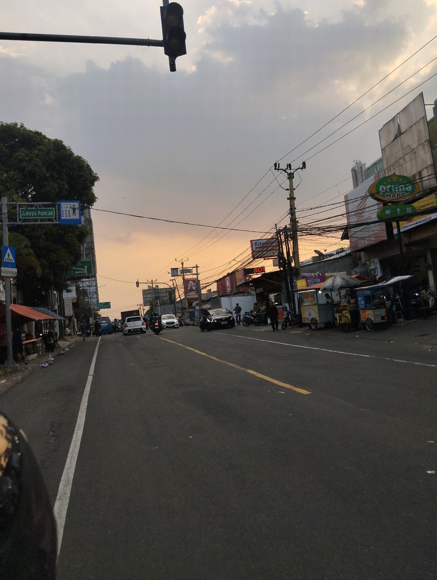 The image depicts a street scene in Megamendung, Indonesia, during what appears to be dusk or early evening. The sky is overcast with patches of warm, orange light indicating sunset. The road is asphalt, marked with a white lane divider and a yellow dashed line. On the left side of the road, there are trees, a traffic light suspended from a horizontal pole, and a pedestrian crossing sign. Further down the street, a blue van is visible, along with several cars and motorcycles, suggesting moderate traffic.
The right side of the street features buildings with various signage, including a prominent green and yellow sign for "Prima MART" and another for "Alfamart". Several small food stalls or carts are set up along the sidewalk, some with umbrellas providing shade, and people are seen near these stalls. Electrical wires are strung across utility poles, indicating the presence of infrastructure. The overall atmosphere is that of a busy, functional urban environment transitioning into nighttime.