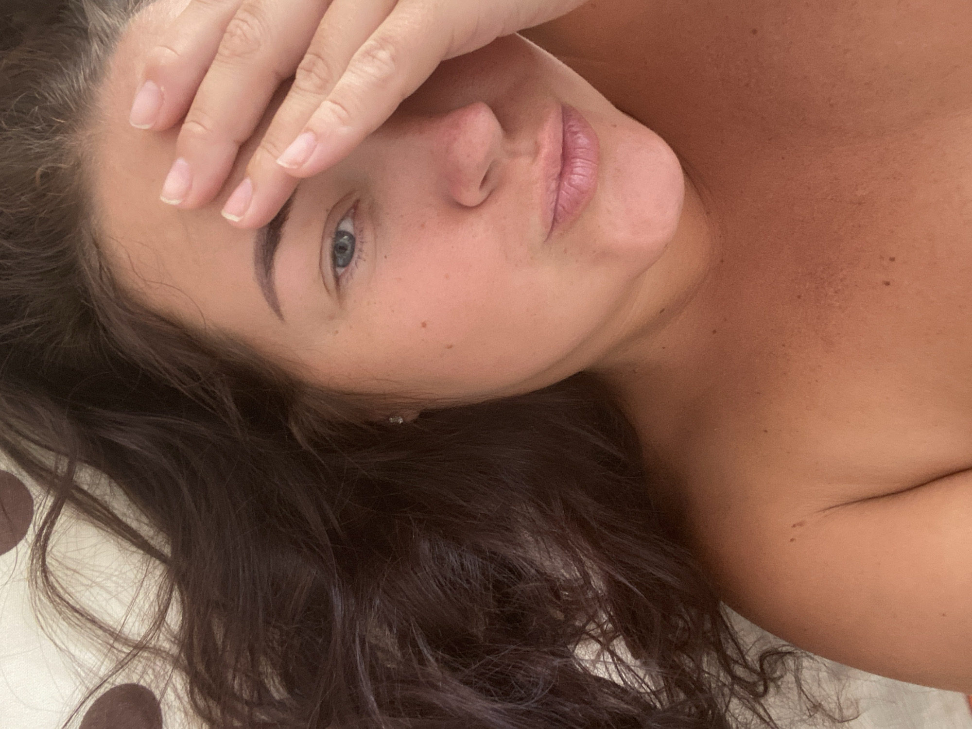 A close-up, over-the-shoulder shot shows a woman with dark, wavy hair covering her right eye and forehead. She is wearing no top, exposing her shoulders and upper chest. Her skin is tanned, with several moles visible. Her lips are slightly parted, and she appears to be looking towards the camera. The background is a patterned fabric, possibly a bedsheet. The image was taken in Sochi, Russia.