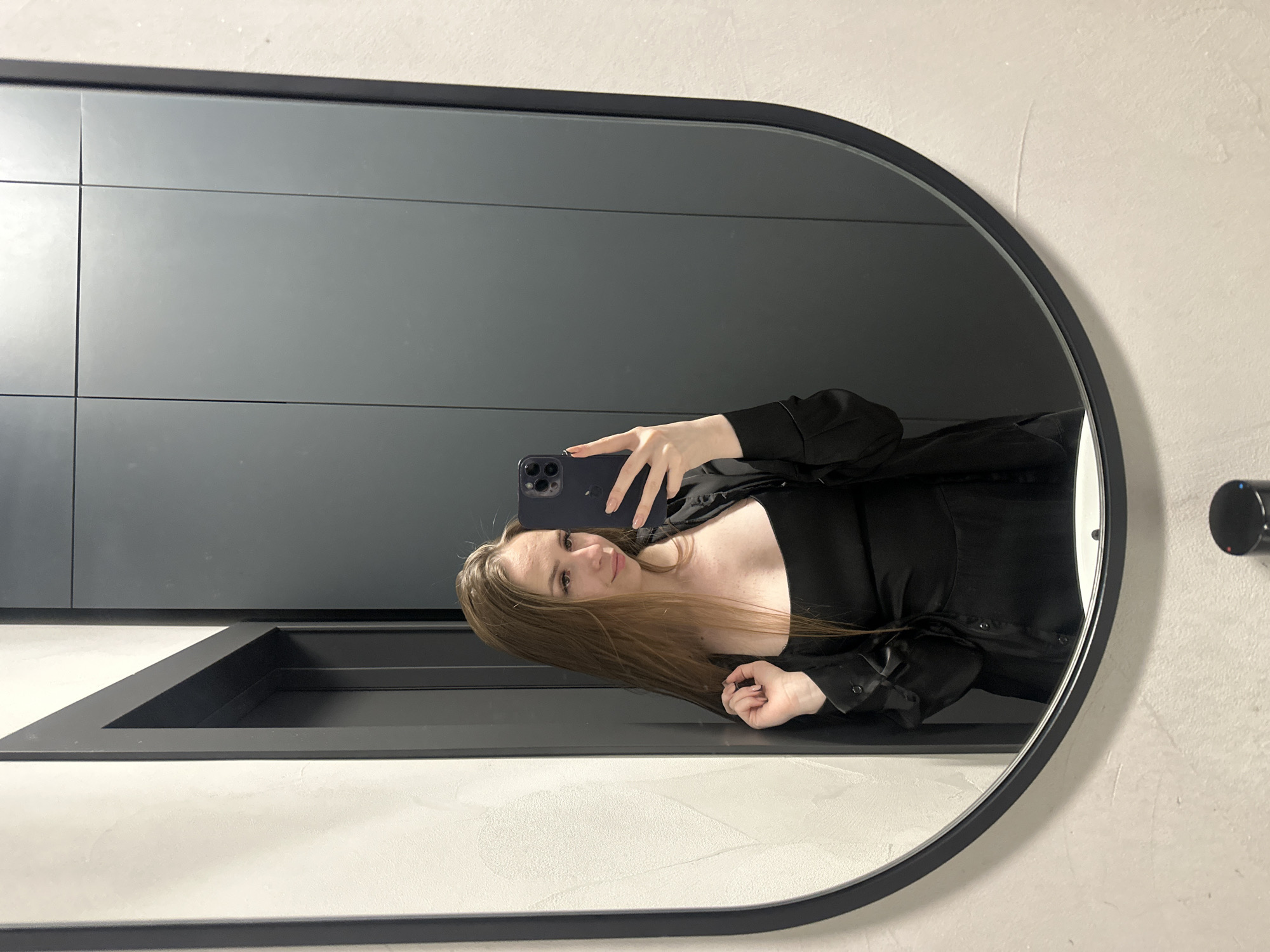 A woman with long, brown hair takes a mirror selfie. She is wearing a black dress and a black open button-down shirt. She is leaning against a textured white wall on the left and her reflection is visible in a large, oval mirror. Behind her reflection, dark gray cabinets are visible. The setting appears to be an interior space with modern finishes, possibly a bathroom or dressing area.