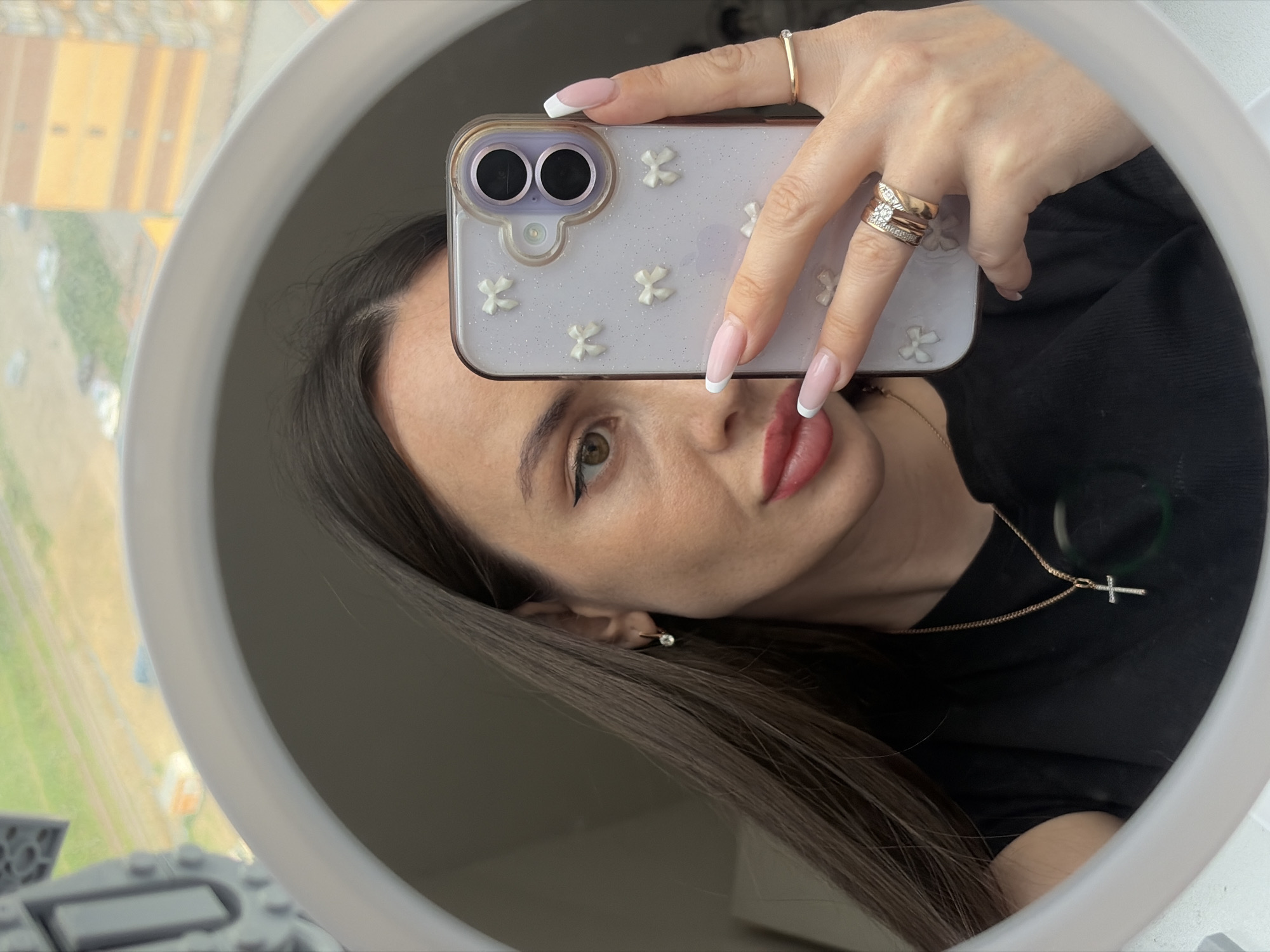 A selfie photograph taken in Saint Petersburg, Russia, shows a woman in a mirror reflection. She is holding a phone with a light-colored case decorated with small, white floral appliques and has manicured nails with a French tip design. The woman has long, dark hair, light skin, and is wearing a black top. She has on a gold necklace with a cross pendant, a stud earring, and several rings on her fingers. In the blurred background visible through the window is an urban scene with buildings.