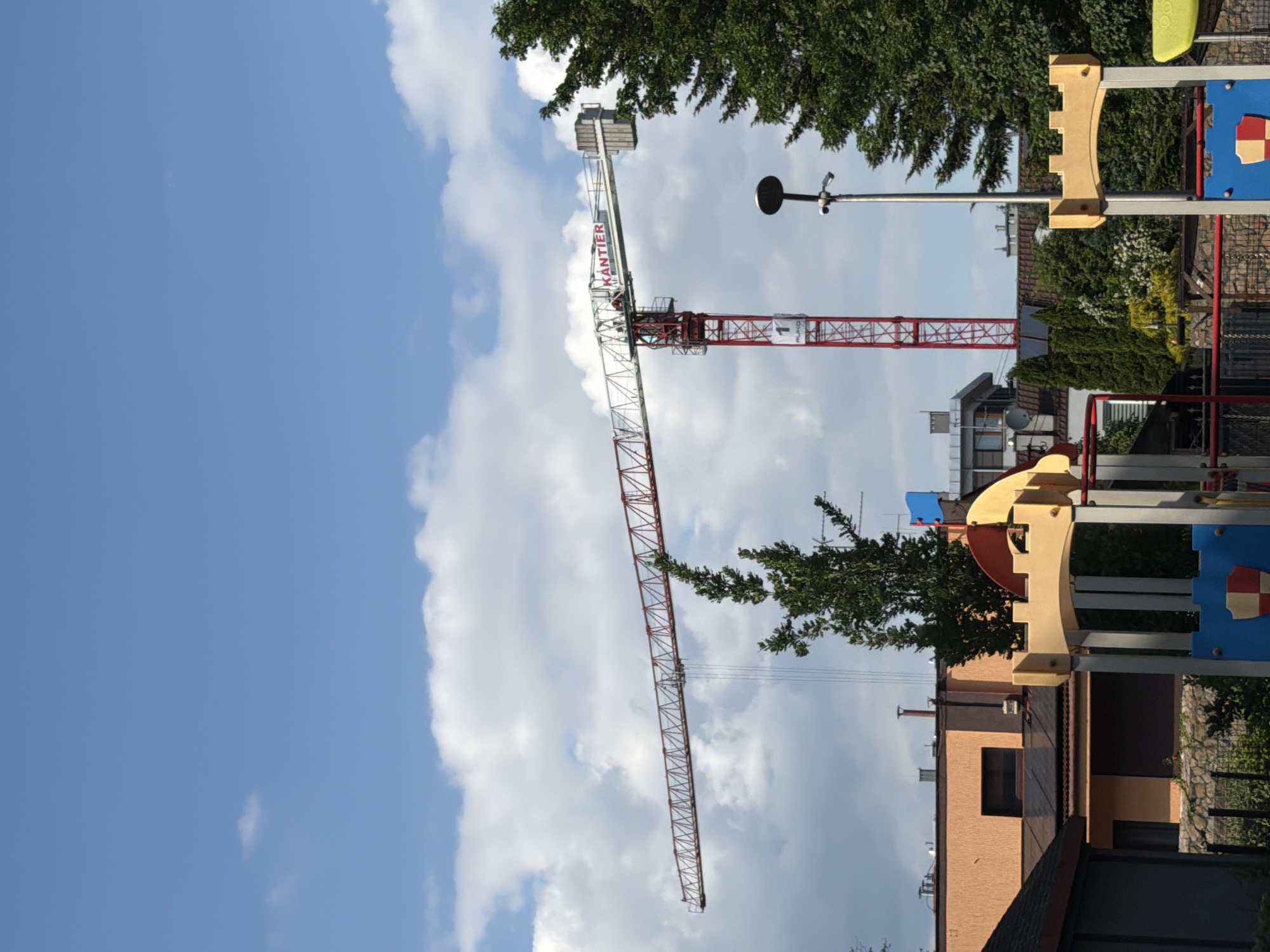An eye-level, medium shot captures a construction crane against a bright blue sky with scattered white clouds, in an urban outdoor setting. The crane's boom extends horizontally from right to left, with its red lattice structure visible. The tower of the crane, also red, rises vertically on the right side of the frame. The word "KÄNTIER" is prominently displayed in red letters on a white sign affixed to the crane's boom.
In the foreground, the tops of buildings and trees are visible. A large evergreen tree is on the right, partially obscuring the crane. Below the crane and trees, there are parts of what appear to be playground structures, with wooden and colored elements. The background features more buildings and trees, suggesting a residential or mixed-use area within the city. The scene is well-lit, indicating daytime.