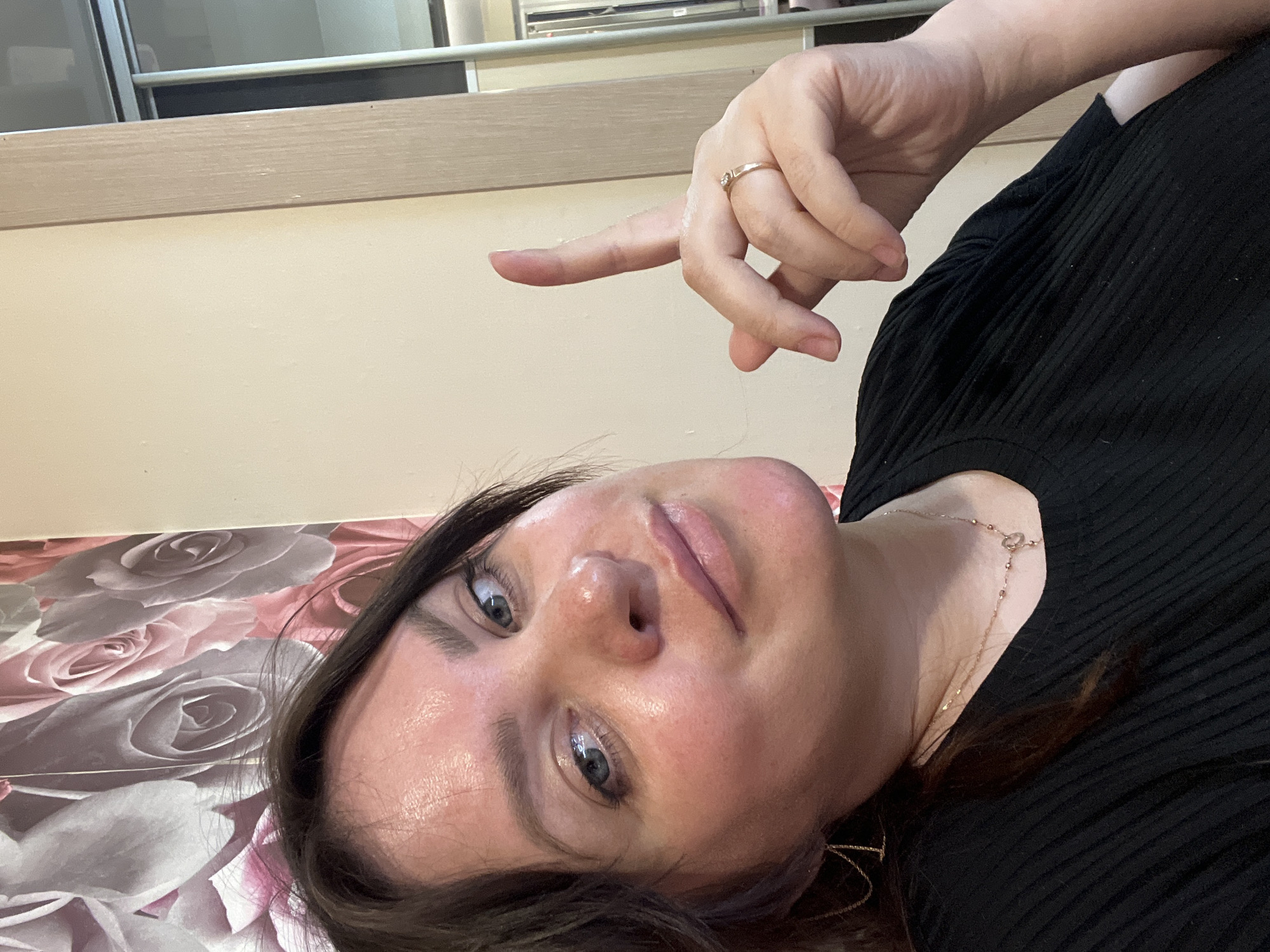 A woman is lying on a surface patterned with roses. She has dark hair and light eyes. Her left hand is raised, with the index finger extended. She is wearing a ring on her right hand and a necklace. The background includes a wall with a light-colored wooden trim and part of a window. The location is not discernible from this image.