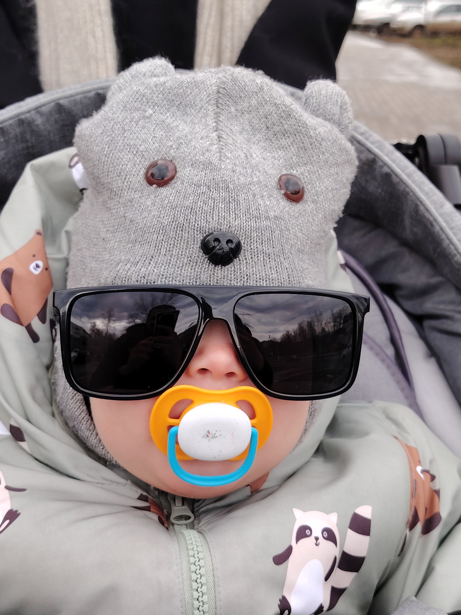 A baby sits in a grey stroller wearing a grey knitted bear hat with brown button eyes and a black button nose, large black sunglasses, and a blue and yellow pacifier. The baby wears a light green jacket with cartoon raccoon prints. The reflection in the sunglasses reveals trees and a cloudy sky. The stroller is partially visible, as is a blurred background that suggests an outdoor setting, possibly in the city of Chaikovsky, Russia.