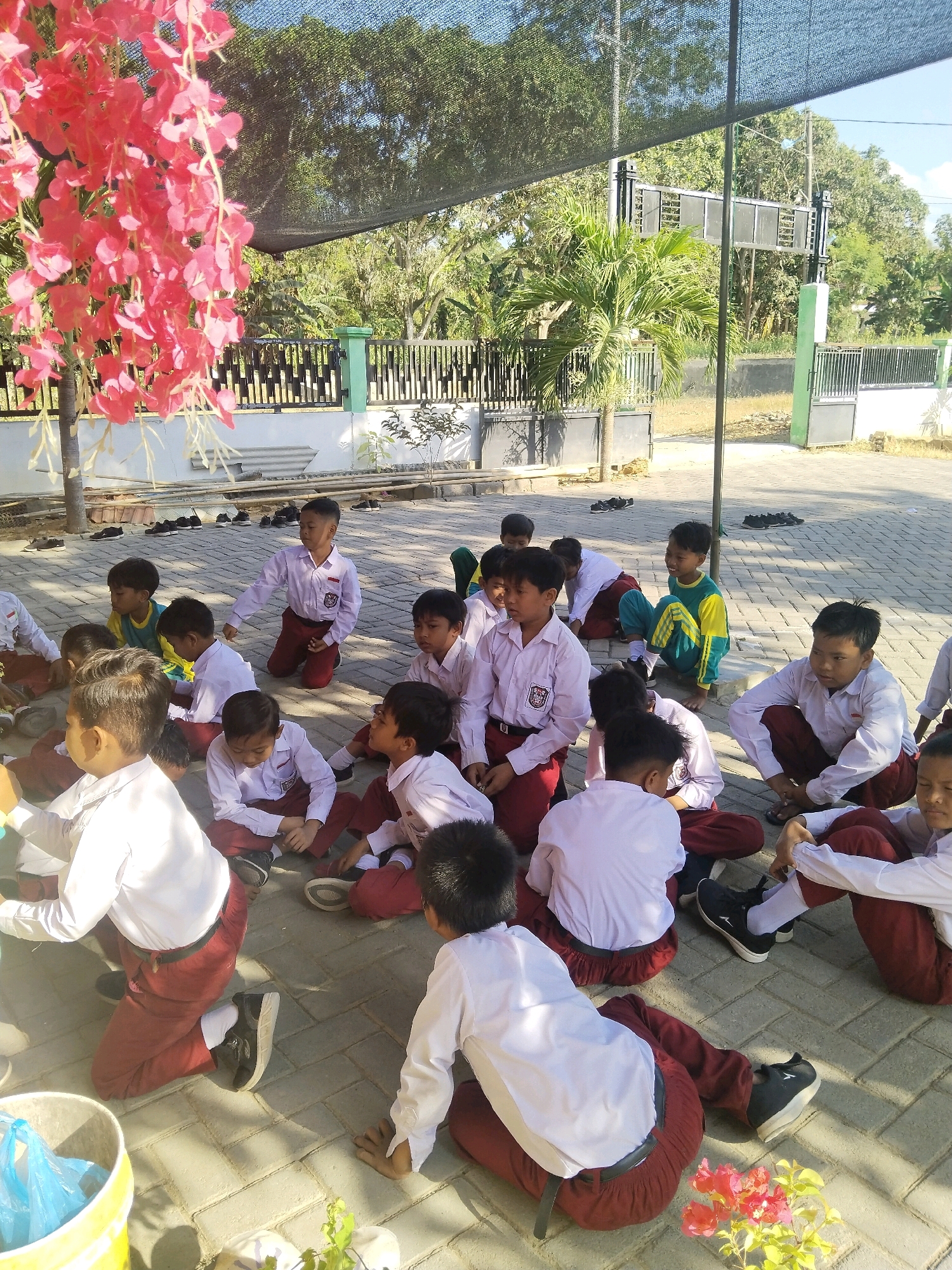 In Taraban, Indonesia, a group of young students, likely elementary school-aged, are gathered outdoors in a paved courtyard. They are all dressed in white long-sleeved shirts and maroon pants, suggesting a school uniform. The students are sitting or kneeling on the ground, some facing the camera and others turned away, in what appears to be a classroom or lesson session.
The environment includes a fence in the background, with trees and foliage visible beyond it. A black mesh shade structure is stretched overhead, providing partial sun protection. Some shoes are neatly arranged on the ground near the fence. In the foreground, a yellow bucket with a blue plastic bag inside is visible, along with a small potted plant and some pink flowers. The lighting suggests it is daytime.