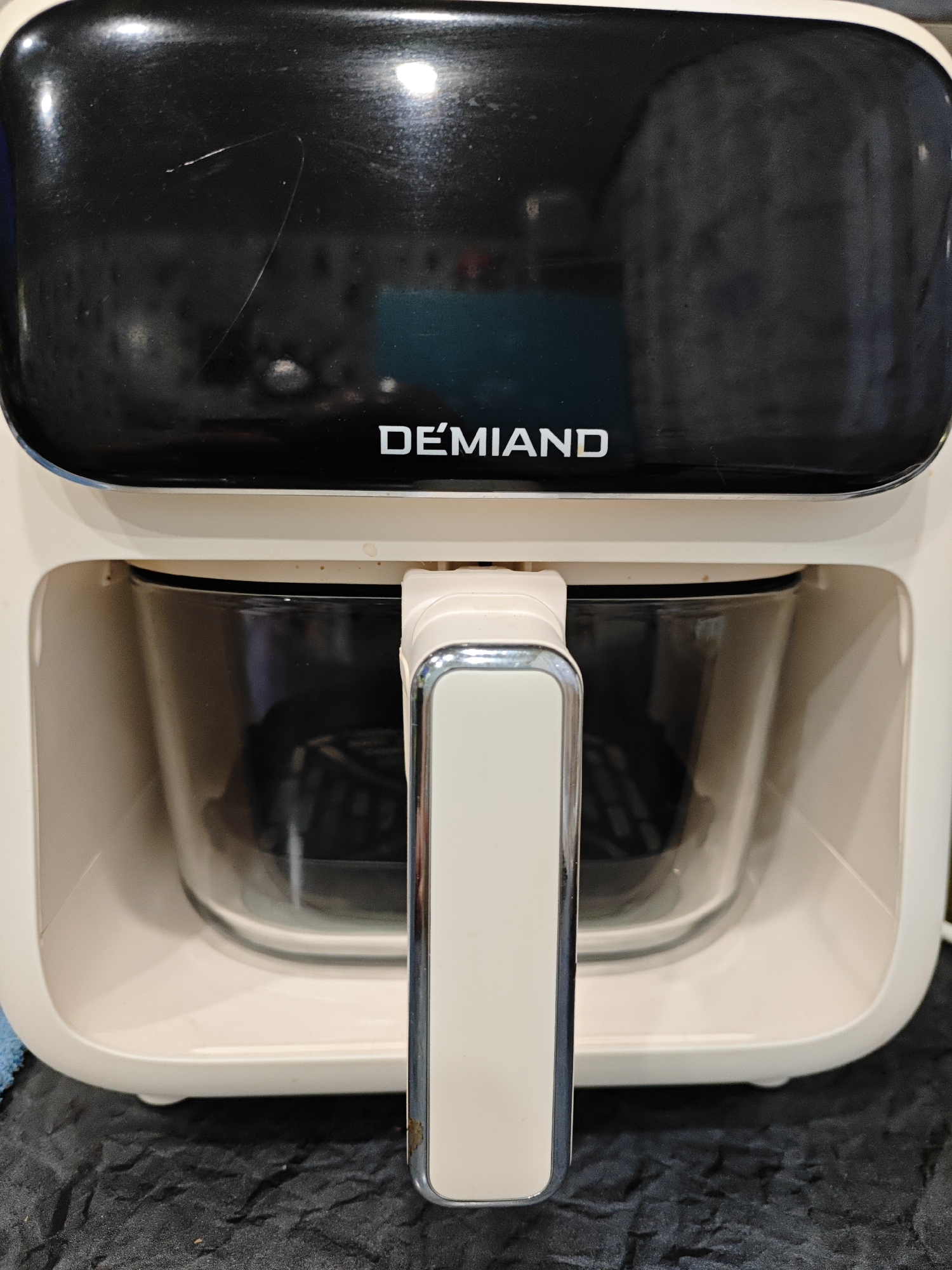 The image shows a close-up of a cream-colored air fryer with a black top. The brand name "DEMIAND" is visible in white lettering on the black top. A clear glass basket with a black interior basket is visible through the front opening of the air fryer. A silver and cream-colored handle extends from the front of the air fryer, in front of the glass basket. The air fryer is placed on a black, textured surface. There are no people or any other objects visible in the frame. The location is not specified in the image.