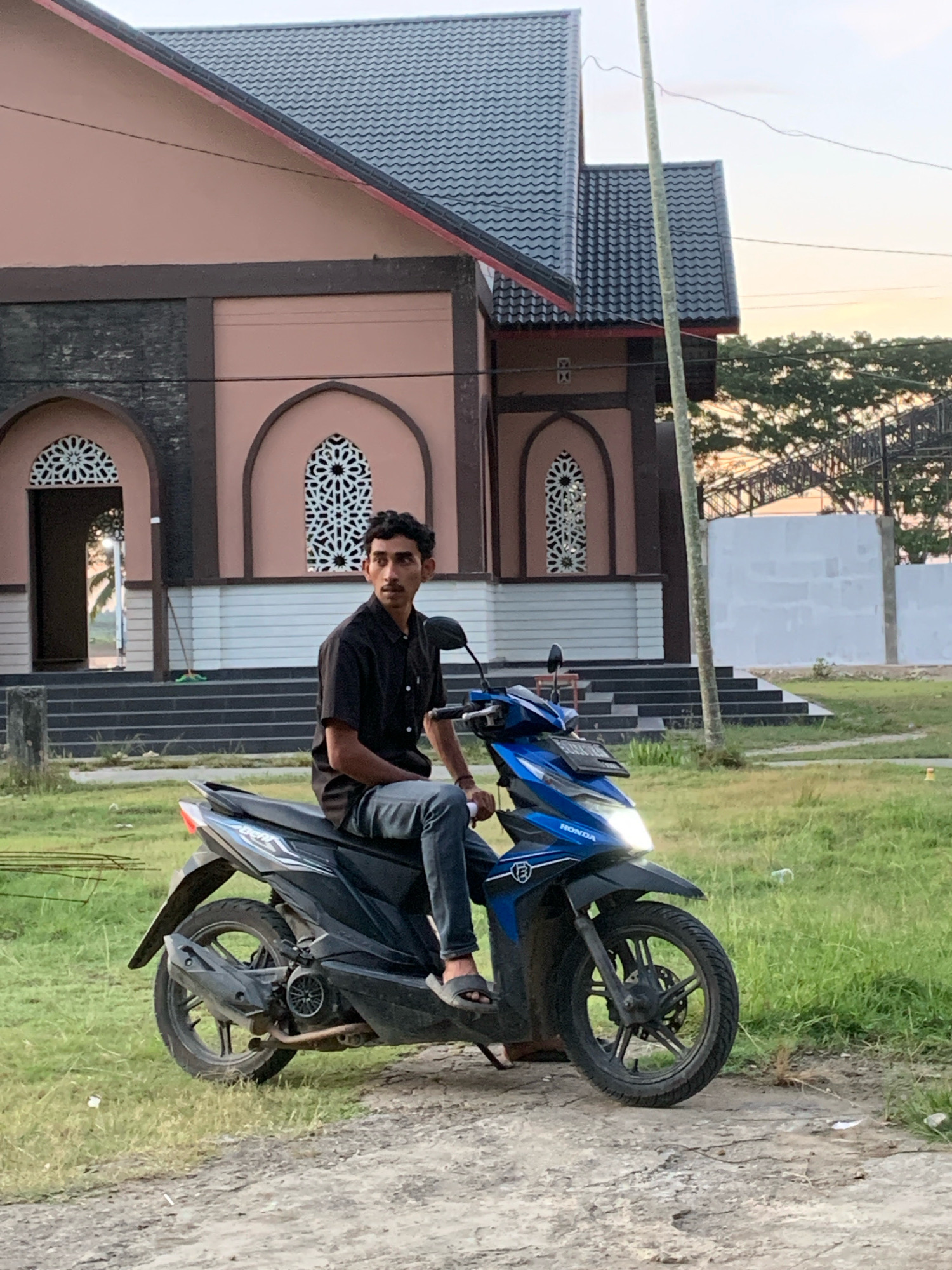 A man wearing a black button-down shirt and dark jeans is sitting on a blue Honda motorcycle on a dirt path. He is looking to his right, with his left hand on the handlebar and his right hand resting on his leg. The motorcycle has its headlight on. Behind the man and motorcycle is a grassy area with a building in the background. The building is pink with a dark grey tiled roof and arched windows with decorative white latticework. Steps lead up to the building. The sky is a light orange, indicating either sunrise or sunset. The scene appears to be in a rural or suburban area of Palohawe, Indonesia.