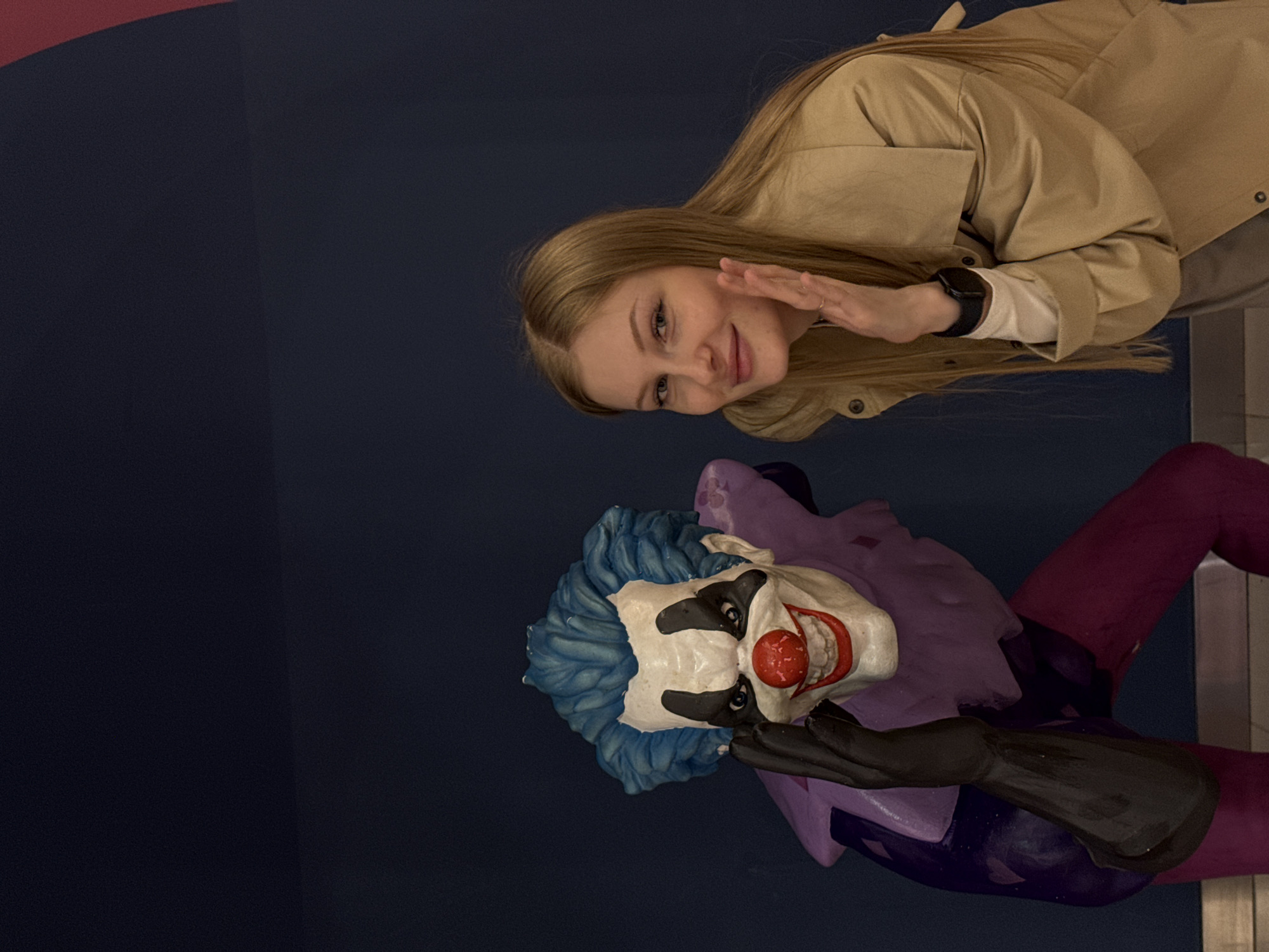 A fair-skinned, blonde woman in a tan jacket poses with a clown statue. The statue is a painted figure of a clown with blue hair, white and black face makeup, a red nose, a purple collar, and a purple and dark purple body with dark sleeves. The background is a deep blue wall. The location is Perm, Russia.