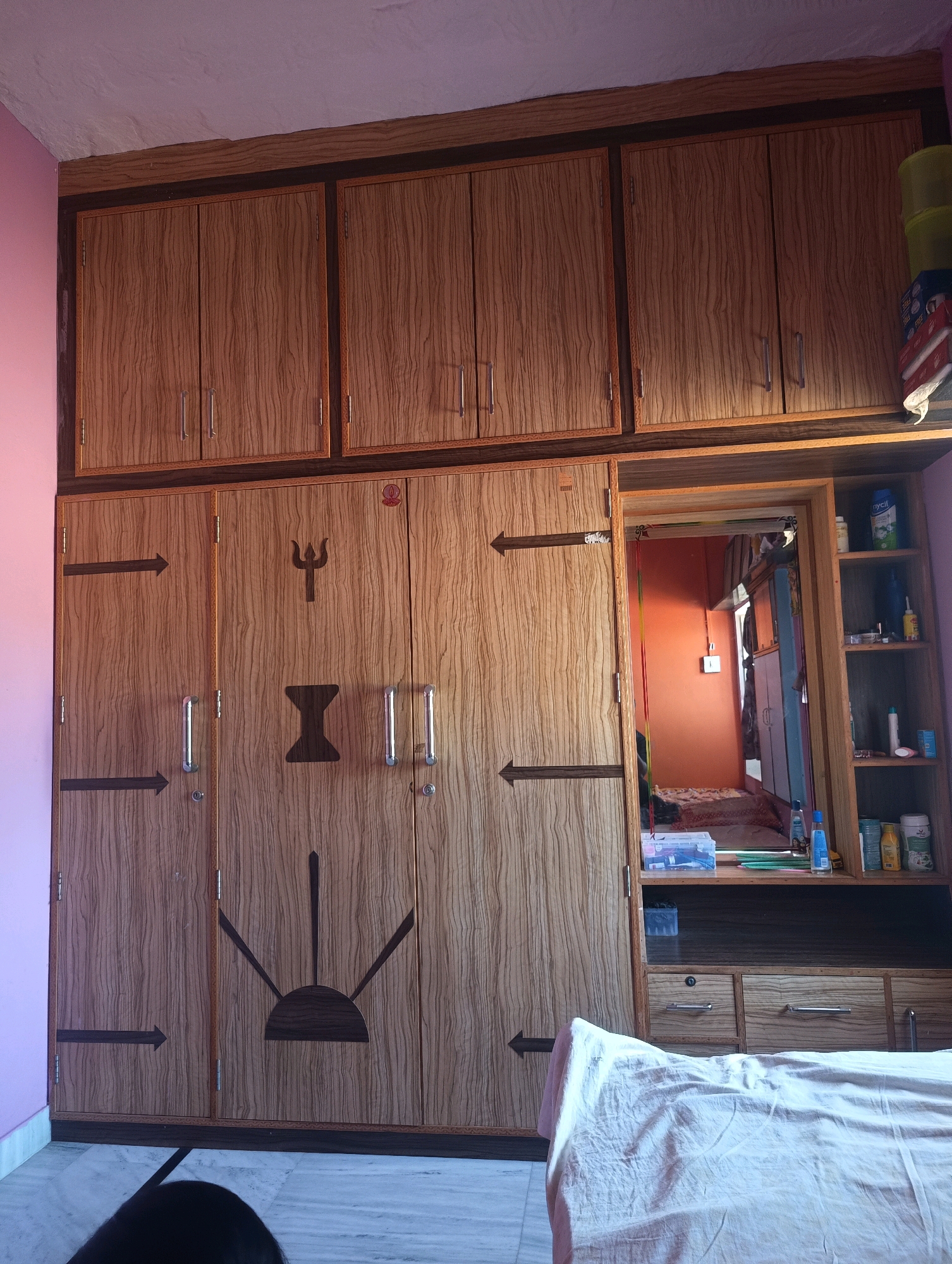The image captures an interior scene of a room, likely a bedroom or dressing area, featuring a large wooden wardrobe. The wardrobe has a wood-grain finish and is divided into multiple sections, with several doors. Decorative motifs are present on some of the doors, including a trident symbol and other abstract designs. Above the main wardrobe, there is a smaller set of cabinets.
To the right of the main wardrobe, a mirrored section is integrated, revealing a glimpse of another room with an orange-colored wall and some furniture. Adjacent to this mirrored section is an open shelving unit containing various bottles, containers, and possibly toiletries or cleaning supplies. The lower part of the wardrobe has drawers with metal handles.
The floor is tiled with a marble-like pattern. A white sheet or bedspread covers a surface in the foreground, partially obscuring the lower section of the wardrobe. The lighting suggests an indoor environment, possibly daytime given the natural light reflected in the mirror. The overall impression is that of a functional and lived-in space. The location is specified as Chhapri Bari, India.