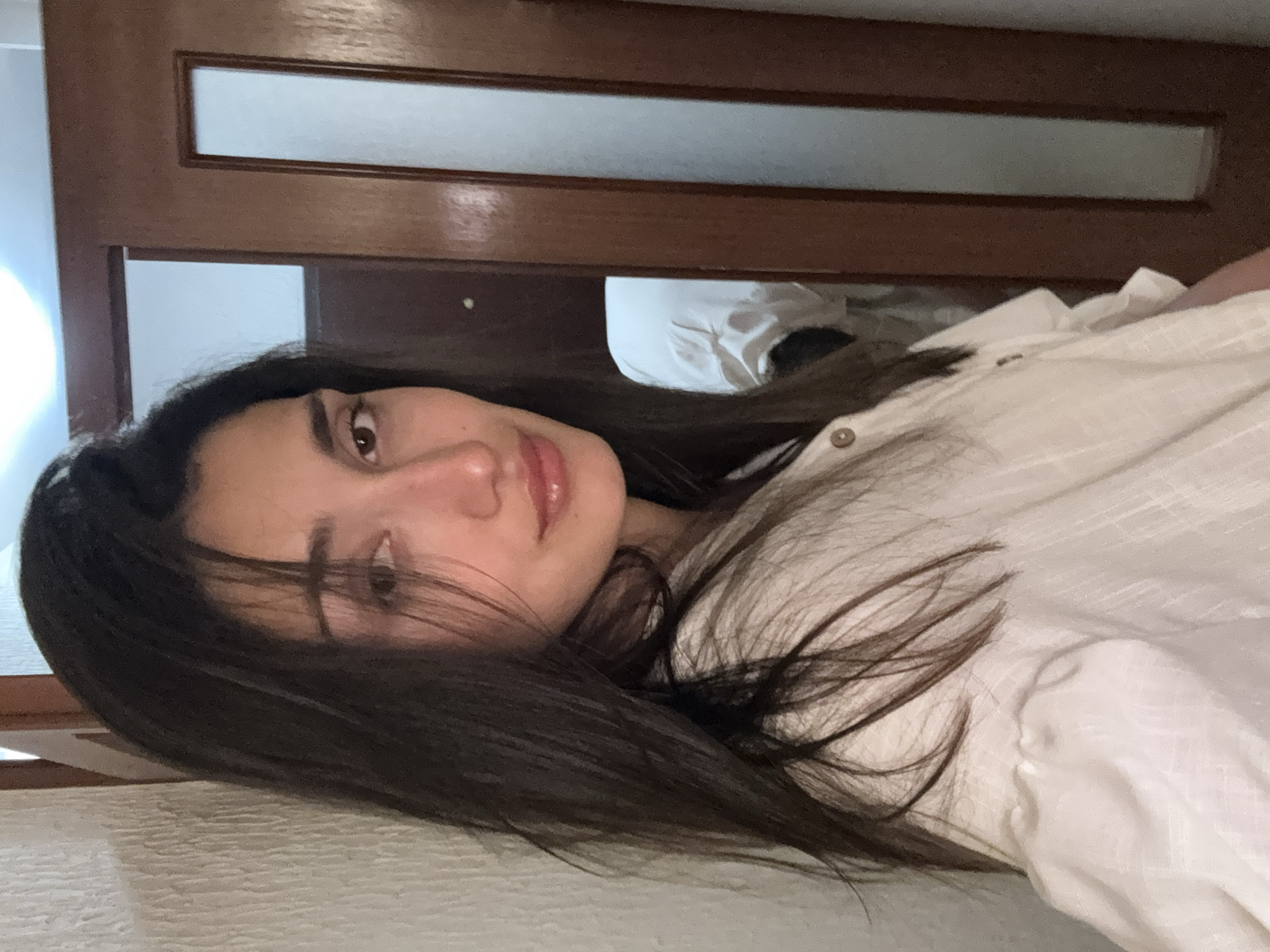 An indoor portrait features a young woman with long, dark hair, lying down and looking directly at the camera. The image is rotated 90 degrees counter-clockwise. She has fair skin, natural eyebrows, and glossy lips, with a soft, relaxed expression. She is wearing a light-colored, possibly white or off-white, collared shirt with buttons.
Her dark hair is spread out around her head and shoulders, blending with the white fabric of her shirt and what appears to be a bed or soft surface she is resting on.
The background consists of a brown wooden door or partition with two horizontal translucent or frosted glass panels, one higher up and another lower down. Behind the lower panel, a lighter wall or surface is visible. The lighting is soft and ambient, suggesting an indoor setting during daytime or early evening. No specific weather conditions or external views are discernible. The overall mood is calm and serene. There is no visible text in the image. The provided location context is Zankovići, Montenegro, but no specific visual cues within the image confirm this location.