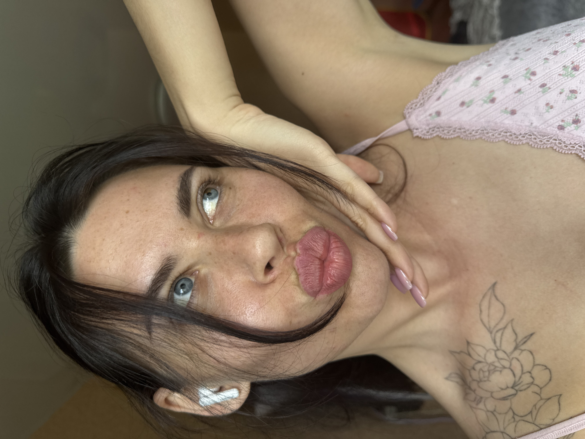 The image shows a close-up of a woman's face and upper body. She has dark hair, blue eyes, and fair skin with freckles. She is wearing white earbuds in her left ear and has her hand on her cheek, with her lips pursed. She is wearing a pink floral lace top. There is a tattoo of a flower on her left shoulder. The background is out of focus and appears to be indoors. The woman is located in Chaikovsky, Russia.