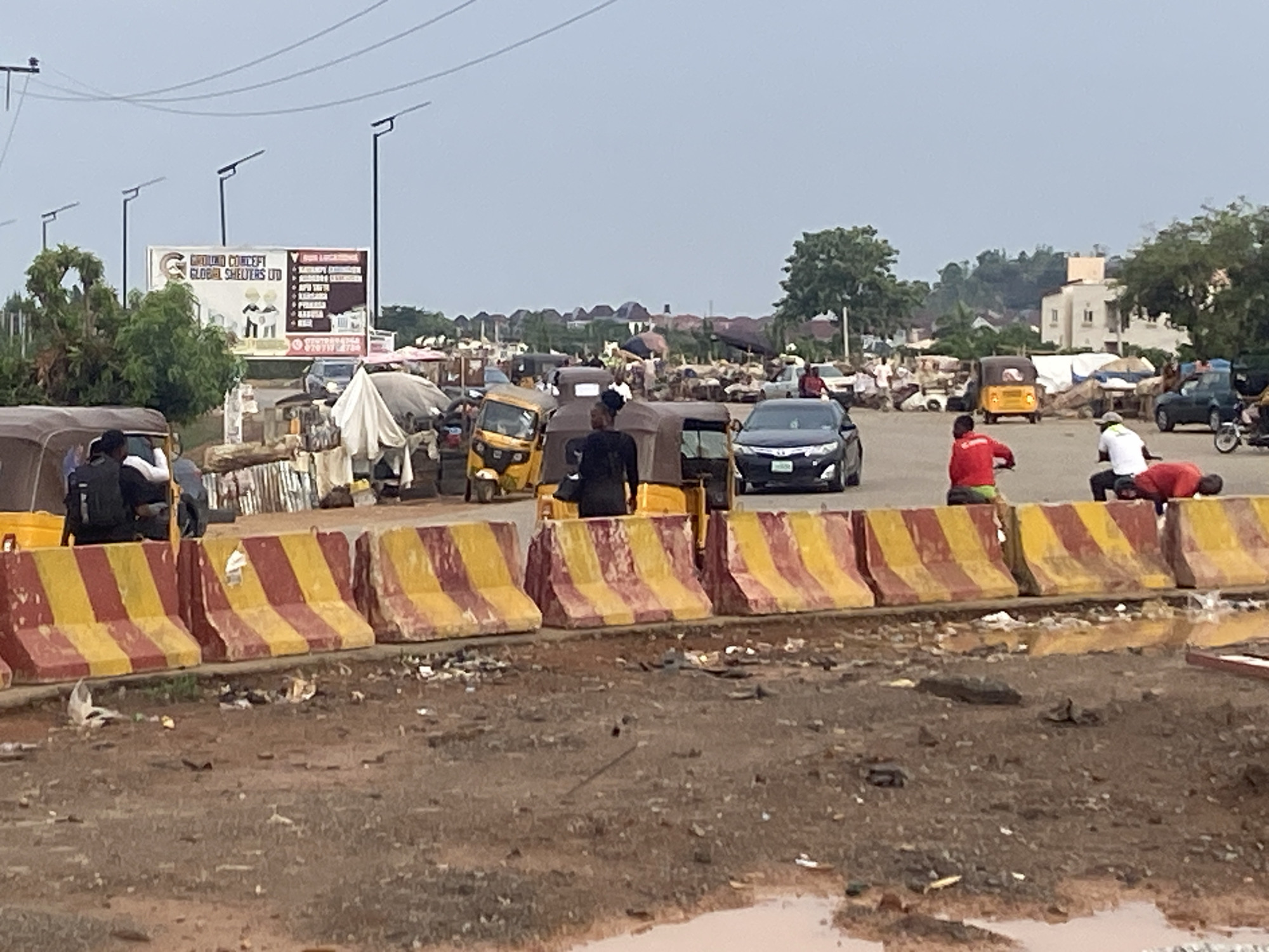 The image shows a bustling roadside scene in Abuja, Nigeria. A row of yellow and red striped concrete barriers separates a muddy foreground with puddles from a congested road. Behind the barriers, several "keke" (auto-rickshaws) and a dark-colored sedan are visible amidst a market scene. People are milling around, some interacting with the vehicles. A billboard is visible in the background, displaying business information. Streetlights line the road. The sky is overcast.