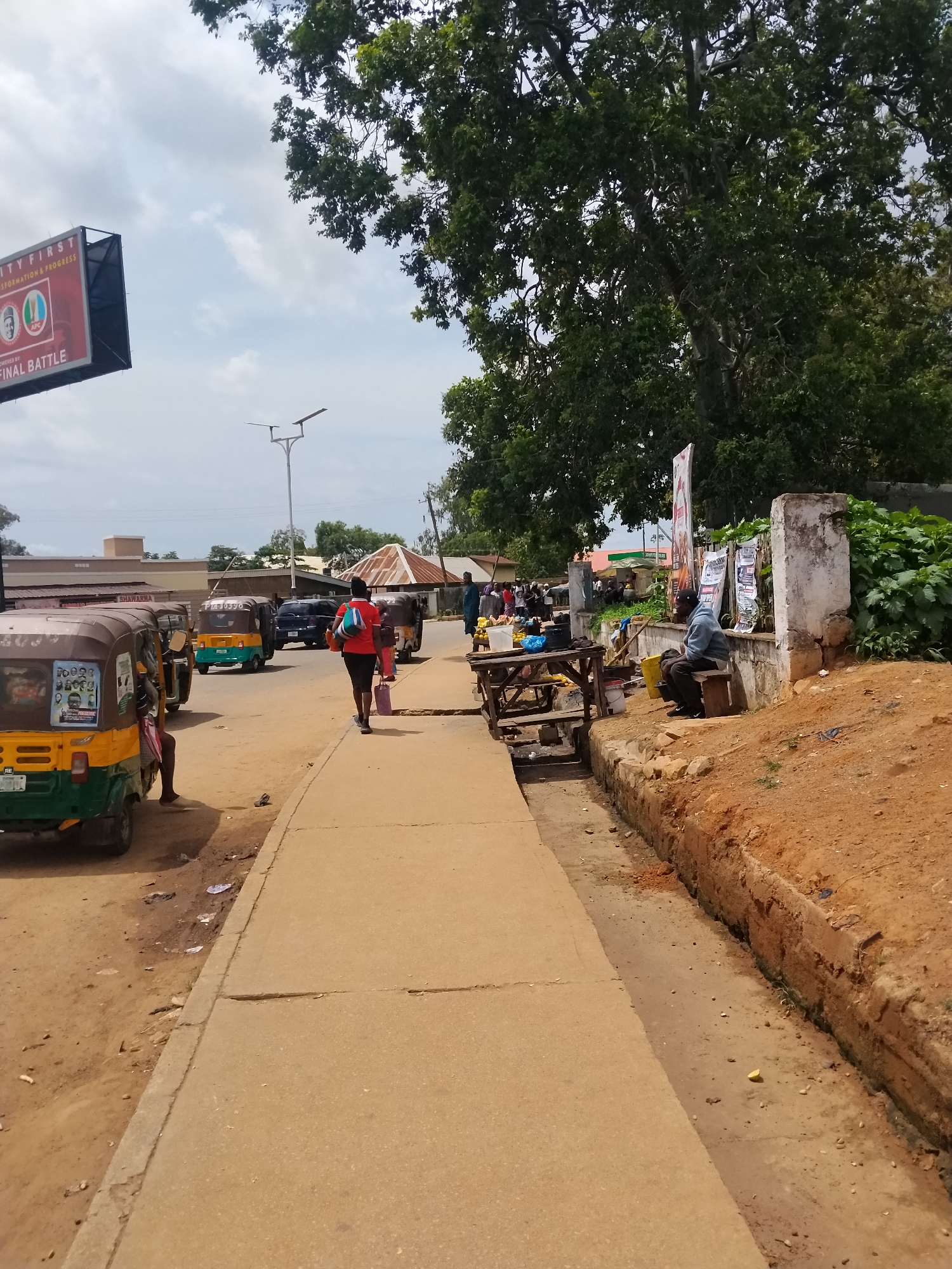 The image depicts a street scene in Jos, Nigeria, on a cloudy day. The foreground shows a concrete sidewalk running alongside a dirt road. Several auto-rickshaws, commonly known as "keke," are parked or moving on the road. One keke in the foreground has posters on its side, featuring portraits of individuals. A person in a red top and black shorts walks away from the camera on the sidewalk, carrying shopping bags. Further down the sidewalk, a man in a blue hooded sweatshirt sits on a wooden bench next to a table laden with items.
The environment includes a large tree with dense foliage in the background, casting some shade. Buildings are visible behind the trees, including one with a peaked roof and another with a flat top. A large billboard with text and images is also present. The overall scene suggests a busy commercial area with ongoing activity.