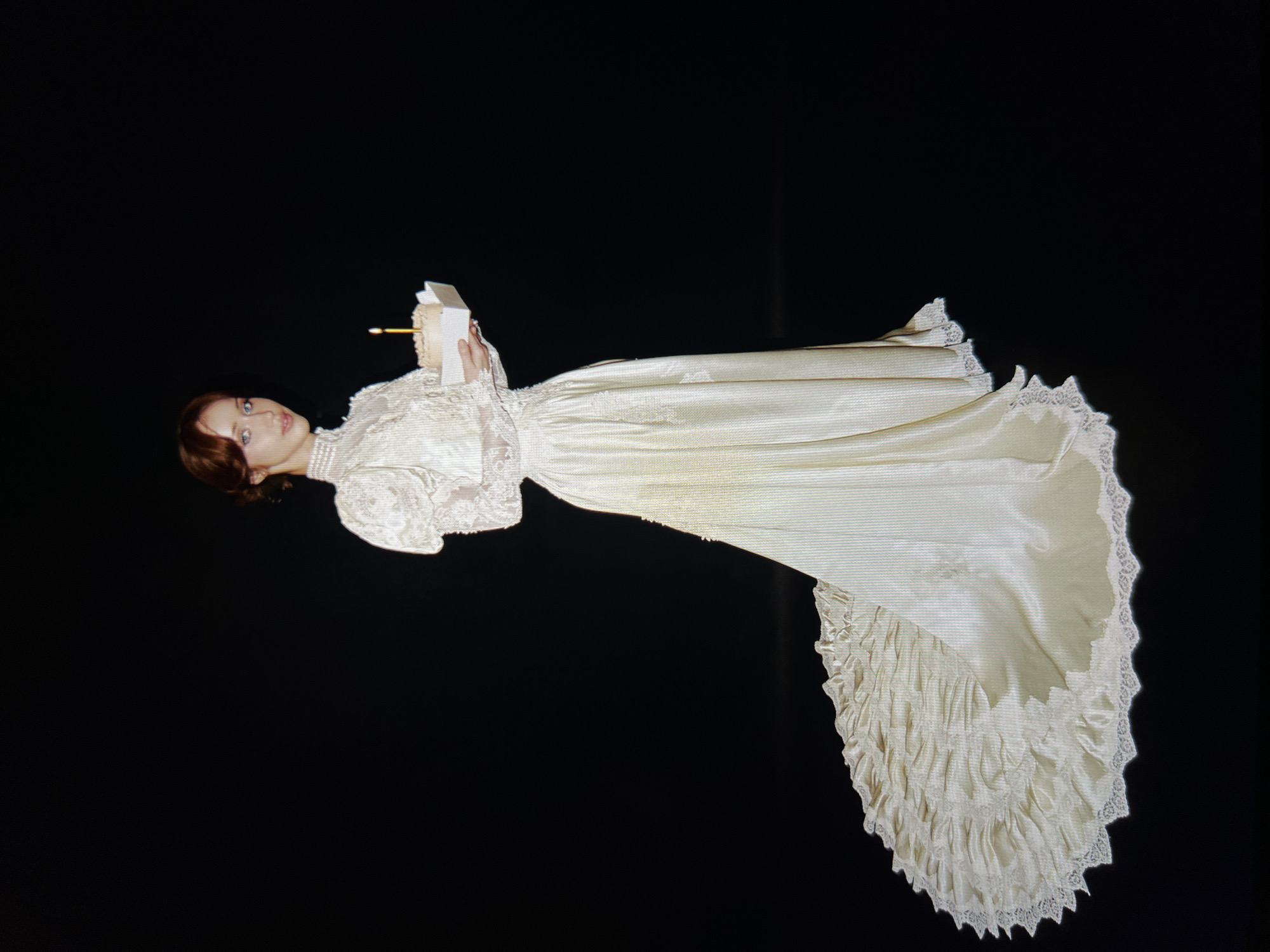 A young woman with auburn hair pulled back and blue eyes stands against a black background, wearing a vintage-style cream-colored gown with a high neckline, puffed sleeves, and a long train. She holds a small, light-colored cake with a single lit candle in her hands. The dress features lace detailing on the sleeves and neckline, and the train is adorned with ruffles and lace trim.