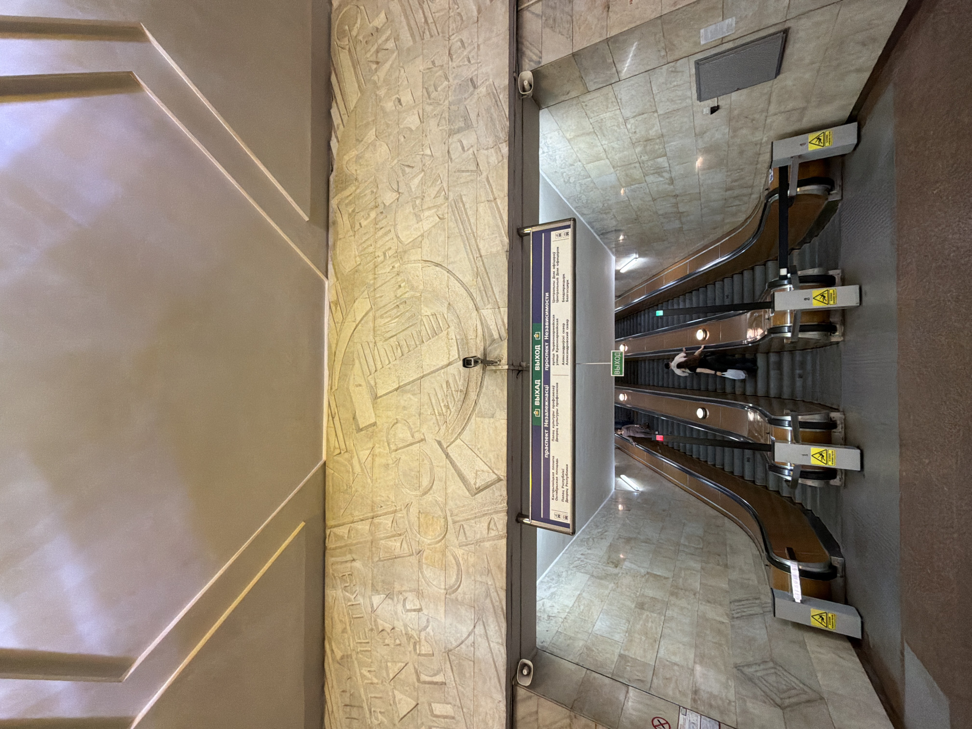 The image shows an entryway to a metro station in Unknown, Unknown. The scene is captured from a high angle, looking down towards a set of escalators. There are two sets of escalators going down into the station. Several people are descending on the escalators. A sign indicates the exits (ВЫХАД and ВЫХОД) to various locations, including the prospekt Niezaležnasci (prospect Nezavisimosti), Kastryčnickaja plošča (October Square), and Palac Respubliki (Palace of the Republic). The walls are decorated with a relief pattern that appears to be Soviet-era artwork depicting USSR. The station has light-colored tile floors and walls.