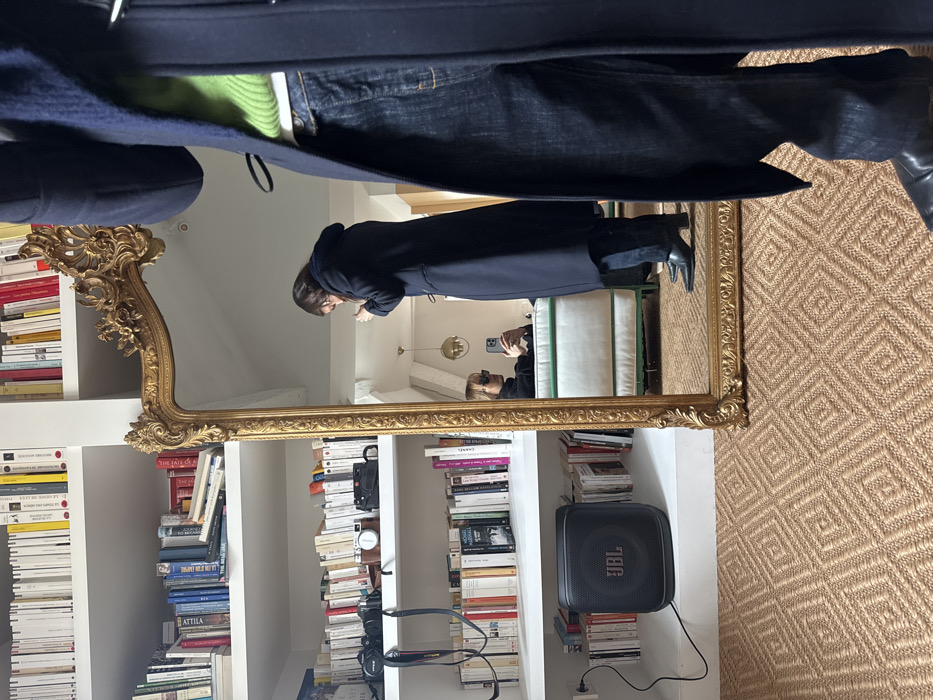 The image depicts an indoor scene within an apartment in Paris, France, captured through a mirror reflection.
On the left side of the frame, a white built-in bookshelf extends from floor to ceiling, densely packed with numerous books, many appearing to be French paperbacks. A black JBL portable speaker is positioned on a lower shelf, connected to a power outlet via a black cable. Also on the shelves are a black SLR camera, a silver rangefinder camera, and a smaller black point-and-shoot camera. The floor is covered with a light brown woven rug featuring a repetitive diamond pattern.
A large, ornate, gold-framed rectangular mirror dominates the center of the image. Partially visible in the immediate foreground, to the right, are the lower torso and legs of the photographer, wearing a navy blue duffle coat over a bright lime green sweater, dark blue jeans, and black pointed-toe boots.
Reflected in the mirror, a person with dark hair, dressed in a long dark navy coat, dark trousers, and black pointed-toe boots, stands in profile, appearing to be adjusting an item near their chest. Further back in the reflection, another individual is seated on a white sofa with a green trim. This seated person wears dark sunglasses and a dark turtleneck top and is holding a white smartphone, seemingly taking a picture or viewing its screen. A brass-colored spherical pendant lamp is also visible in the reflection, hanging from the ceiling.