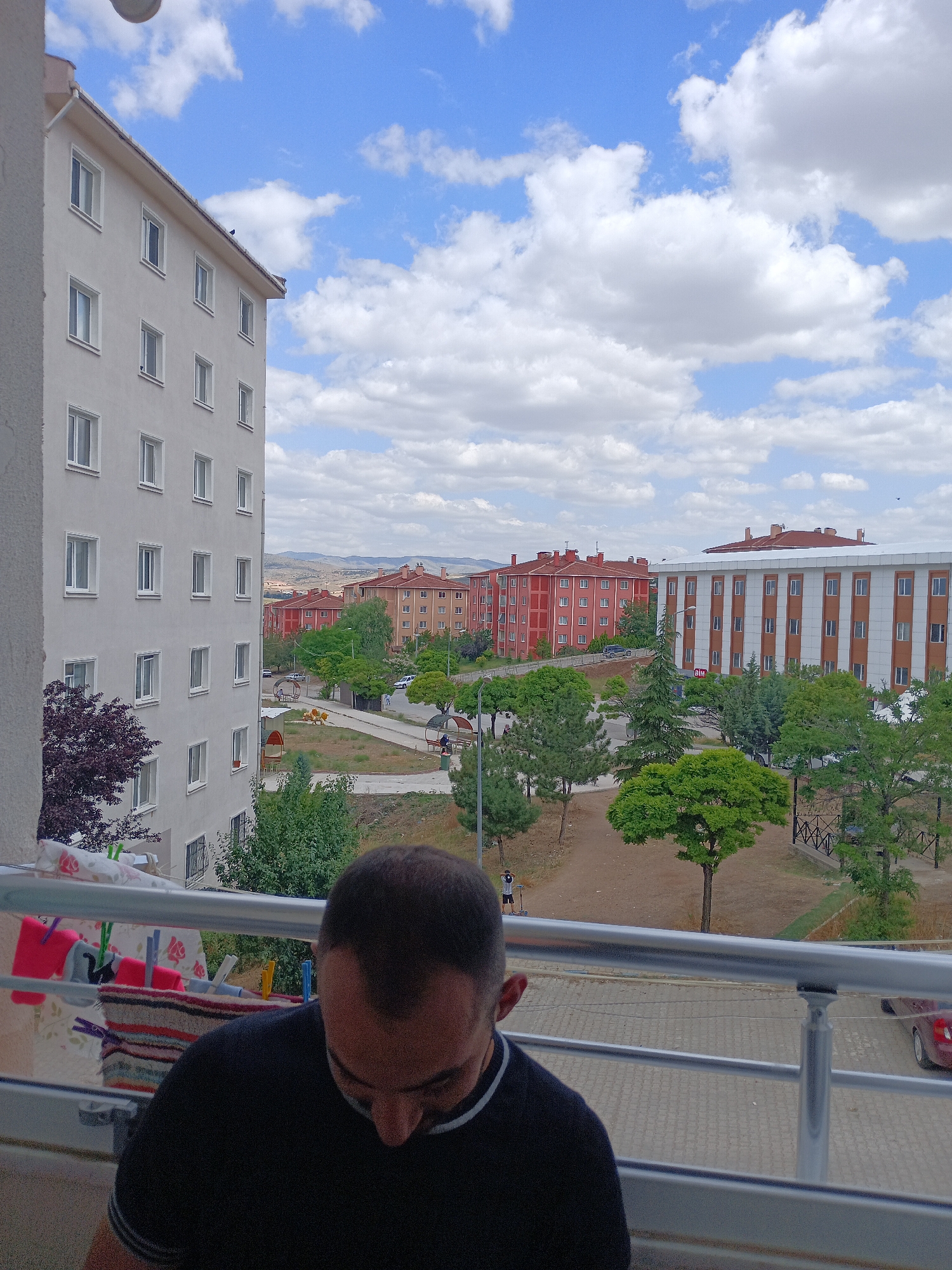 The image captures a view from a balcony in Bahşılı Belediyesi, Turkey. In the foreground, a man wearing a black t-shirt is partially visible, looking downwards. The balcony railing runs across the bottom of the frame. Beyond the balcony, a residential area is depicted with several apartment buildings. The building immediately to the left is a light-colored, multi-story structure with numerous windows. Across from it, there are apartment buildings with red and white facades, and a large white building with orange trim. Green trees and some barren land are visible between the buildings, suggesting a somewhat urban or suburban environment. The sky is blue with scattered white clouds. Clothes are hanging on a clothesline on the left side of the balcony. A red car is parked on the right side of the street. A child on a scooter is visible on a pathway in the middle ground.