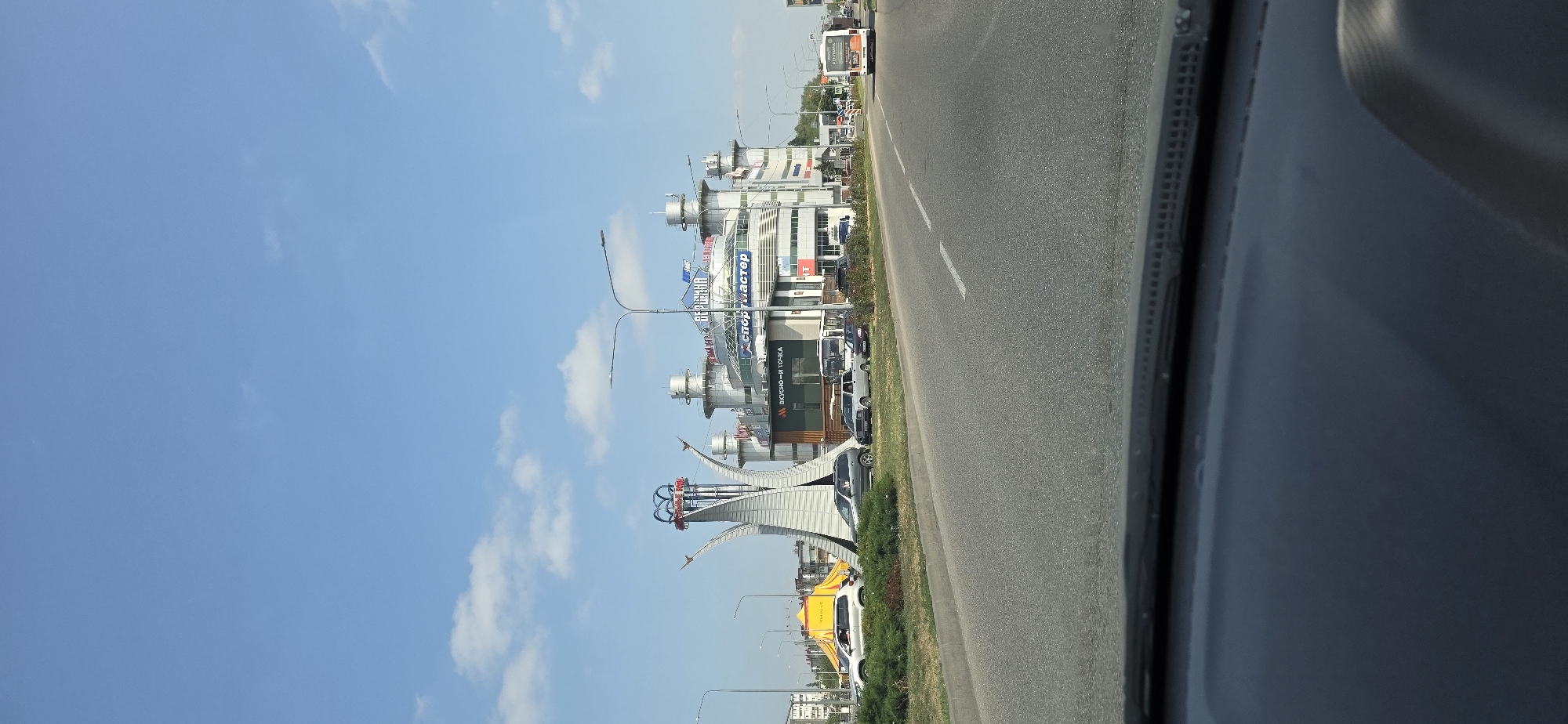 A road view from a car in Mineralnye Vody, Russia, shows a modern, multi-story shopping center with a distinctive architectural design featuring cylindrical towers and a large, stylized metal sculpture in front. The sculpture, resembling an abstract human figure with outstretched arms, stands tall on a grassy median. The shopping center's facade displays signage including "ВЕРШИНА" (Vershina, meaning "Summit"), "СПОРТ-МАСТЕР" (Sportmaster), and "ВКУСНО – И ТОЧКА" (Vkusno - I Tochka, a fast-food chain). A yellow tent is visible to the left of the sculpture. Several cars are parked and driving on the road, and a white bus is seen in the distance on the right side of the frame. The sky is a clear blue with scattered white clouds, and streetlights line the road. The foreground shows the interior of a vehicle, likely a car, with its dashboard visible.