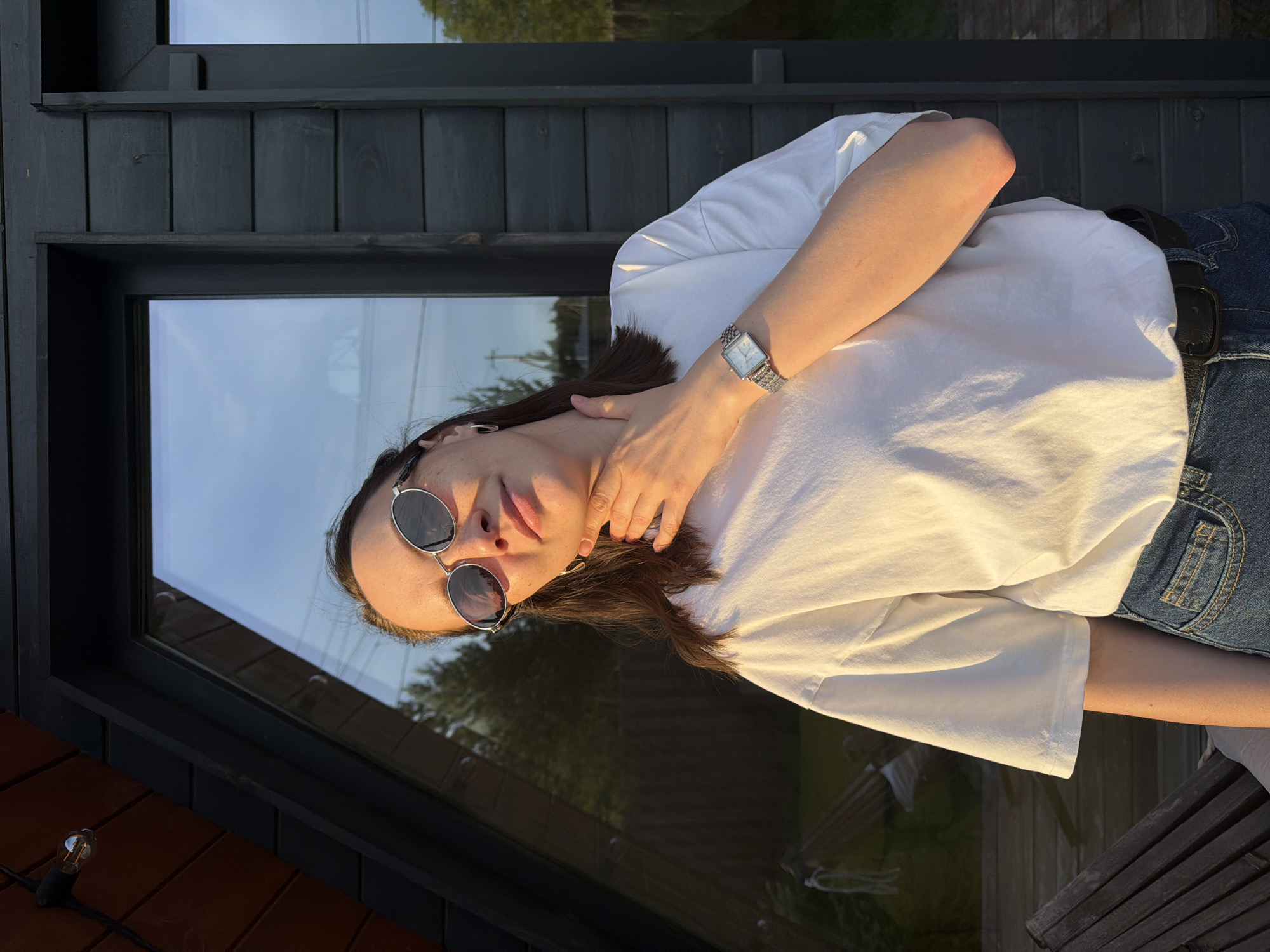 The image depicts a young woman wearing sunglasses, a white t-shirt, and jeans, positioned outdoors near a building with a large window. The light suggests it is either sunrise or sunset. The reflection in the window shows greenery, possibly trees, and sky. The location, Prikamskii, Russia, is not visually verifiable from the image alone.