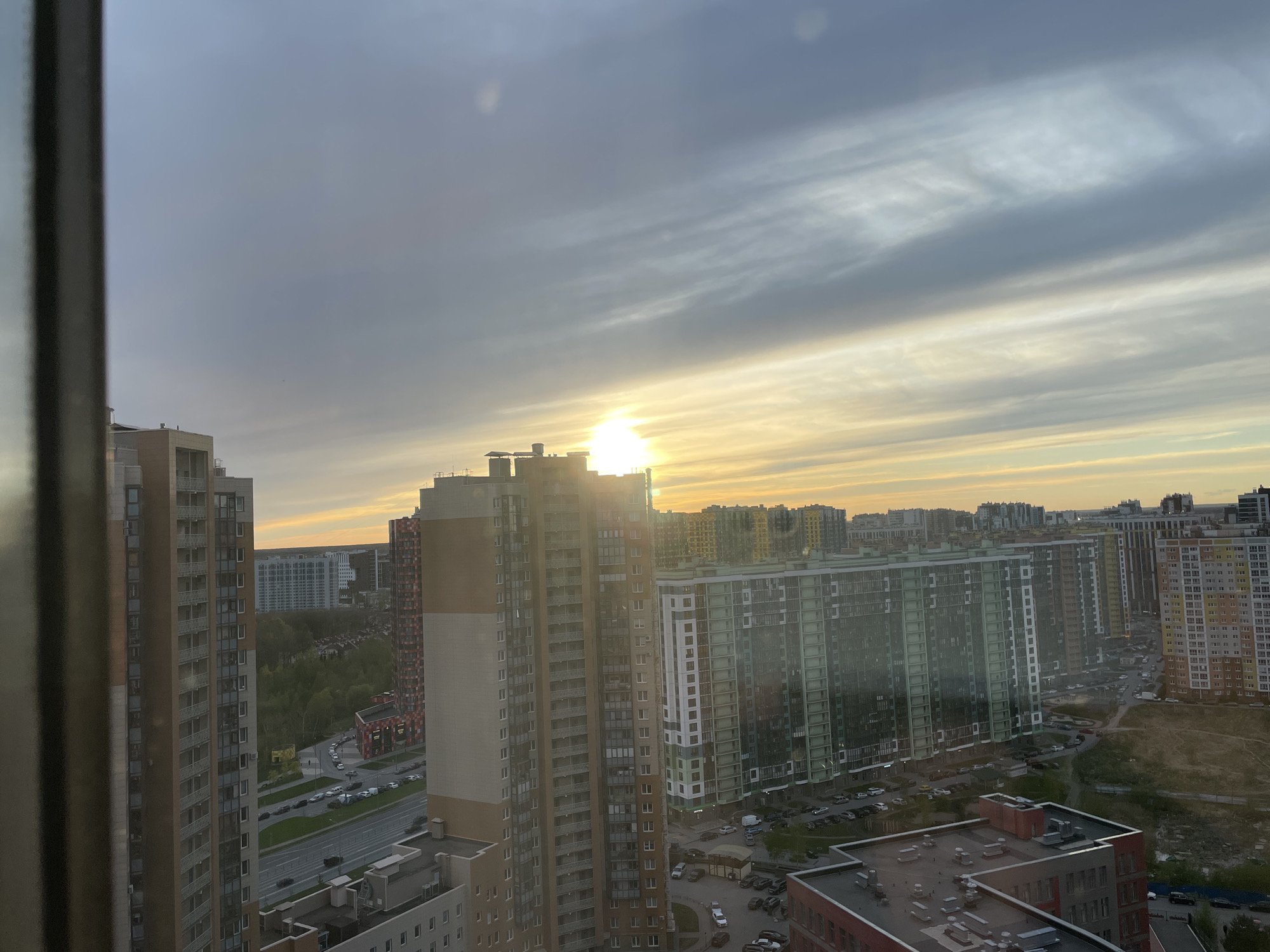 The image shows a cityscape, likely taken from a high vantage point inside a building, possibly in Saint Petersburg, Russia, based on the architectural style of the buildings. The foreground is dominated by apartment buildings of varying heights and designs, constructed of concrete, with varying colors and details. Parking lots and roads with cars are visible among the buildings. The sky is overcast with light breaking through the clouds on the horizon.