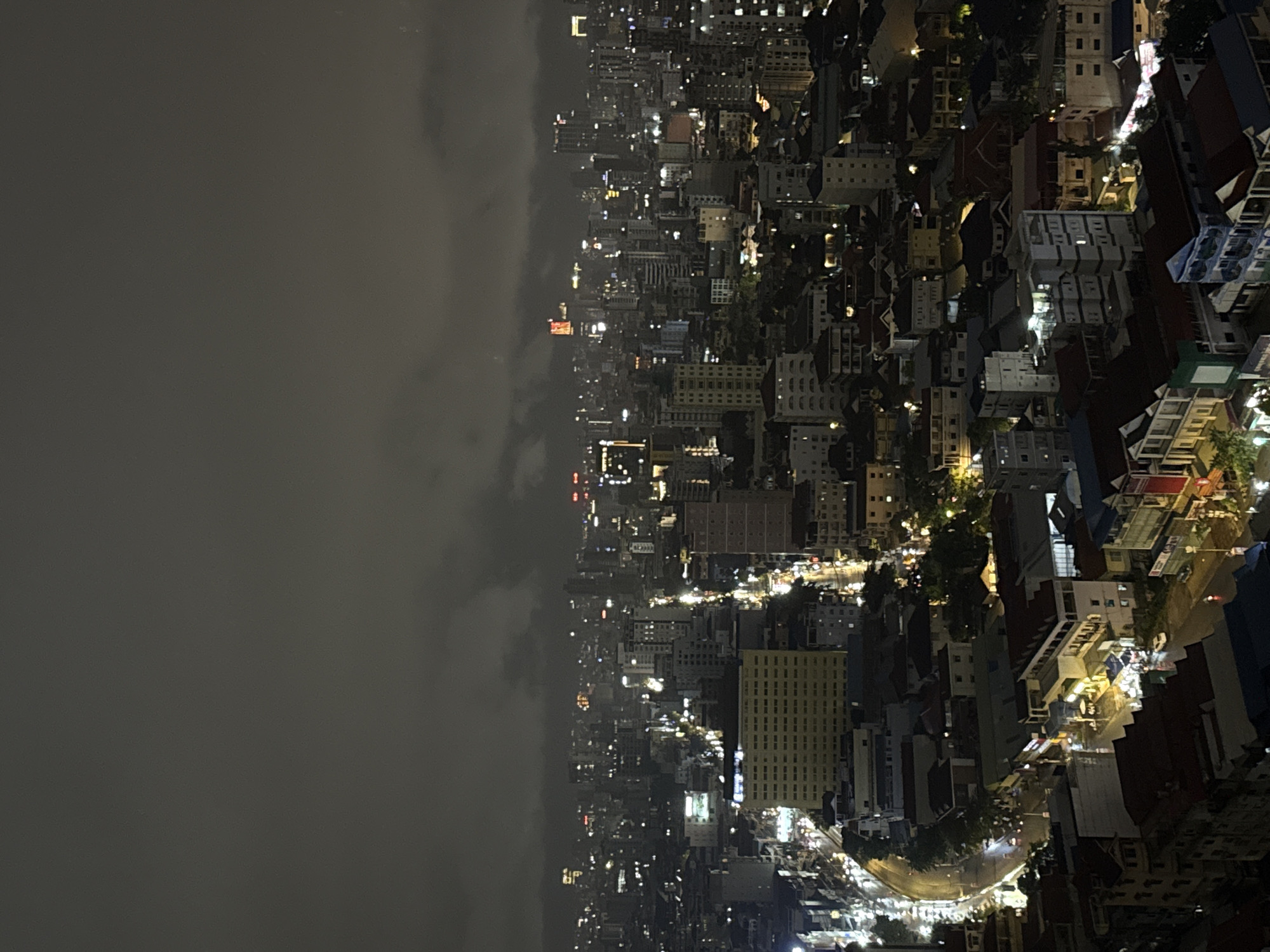 This is an aerial, nighttime photograph of a dense urban cityscape, likely taken from a high vantage point. The scene is dominated by a multitude of buildings of varying heights and architectural styles, illuminated by artificial lights from windows, streetlights, and building facades. The sky is dark and overcast, with some diffused cloud cover obscuring the stars and moon. The city lights create a warm, glowing effect against the dark sky, with a notable concentration of lights forming streets and illuminating individual buildings.
The perspective suggests a location in Khan Mean Chey, Cambodia, a district known for its mixed-use development, including residential areas, commercial establishments, and some industrial zones. The image captures the typical urban sprawl of a developing city, with a mix of modern high-rises and older, lower-rise structures. The street layout appears to be a grid with some curving roads. The presence of streetlights and illuminated vehicles on the roads indicates ongoing activity. The overall impression is one of a bustling metropolis at night, with its own unique character and energy.