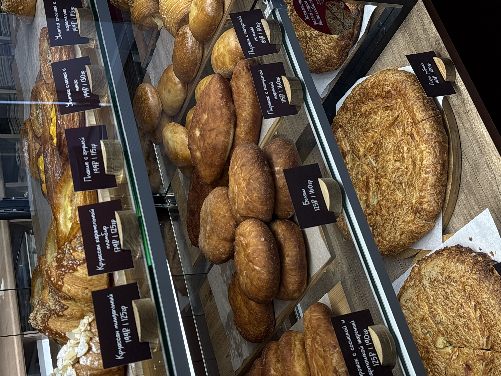A commercial display case presents an assortment of baked goods, likely in a bakery or cafe. The items are arranged in separate sections, each with a label in Russian indicating the type of pastry and its price.
Visible pastries include:
* Varieties of snails with mango and caramel.
* Croissants with almond and caramel.
* Patties with apples
* Patties with cabbage.
* Belashi
* Pirozhki with fried potatoes and sausages.
* Fish cake
The display case has a modern design with a dark frame. The lighting highlights the textures and colors of the baked goods. The case appears to be clean and well-maintained. Based on the writing on the labels, the pastries can be purchased for takeaway.
