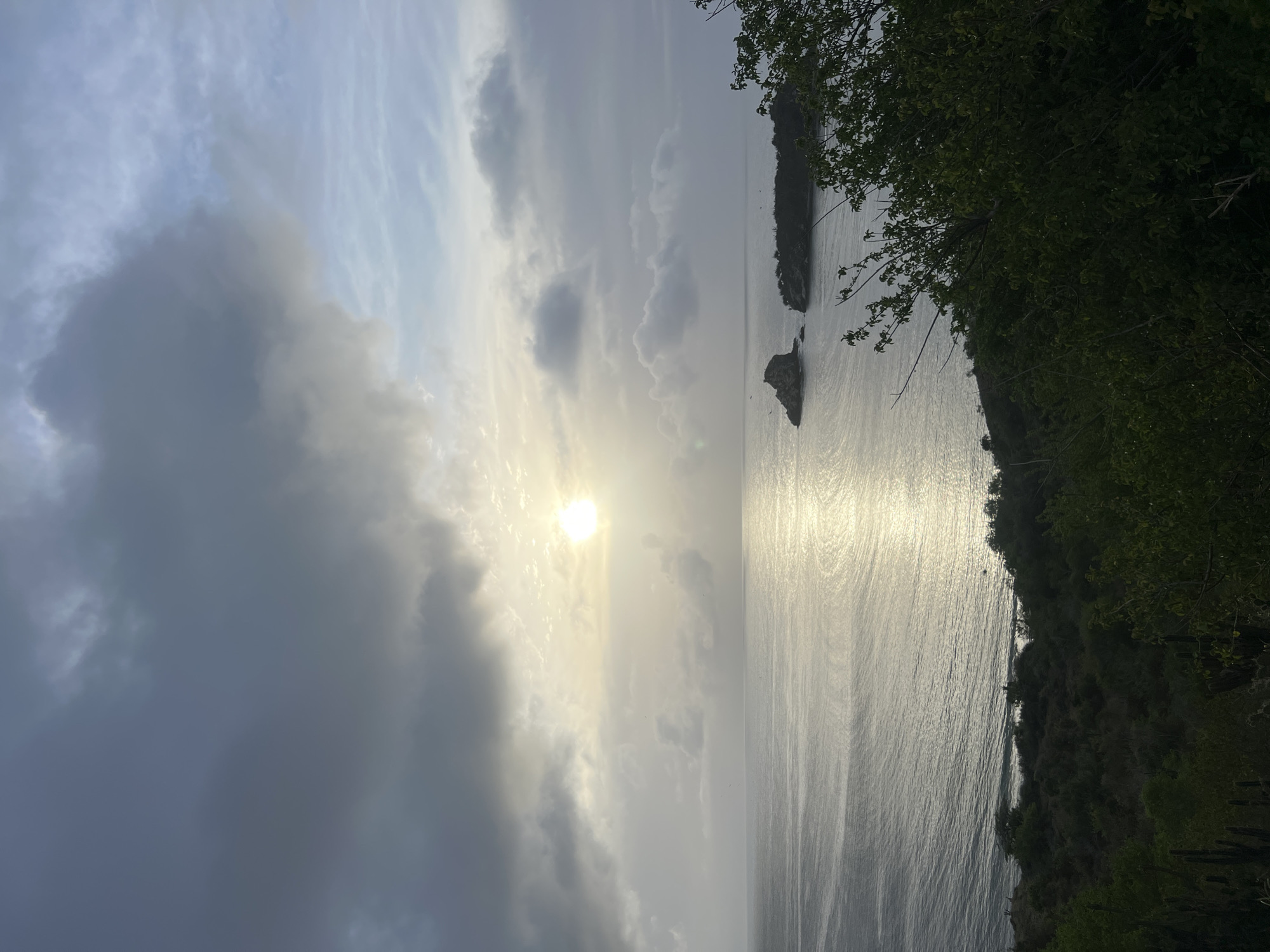 The image depicts a coastal scene under a cloudy sky with the sun partially visible. The sun's rays create a shimmering reflection on the calm sea. In the distance, there are small islands or rock formations jutting out of the water. The foreground is dominated by lush green vegetation, including trees and some cacti. The overall impression is one of a serene natural landscape.
While the image itself does not contain explicit indicators of the city of Manzanillo, Venezuela, this type of coastal environment with islands, tropical vegetation, and a reflective sea is characteristic of many areas along Venezuela's coastline, including regions near Manzanillo. Without further information or landmarks within the image, a precise location within Manzanillo cannot be determined.