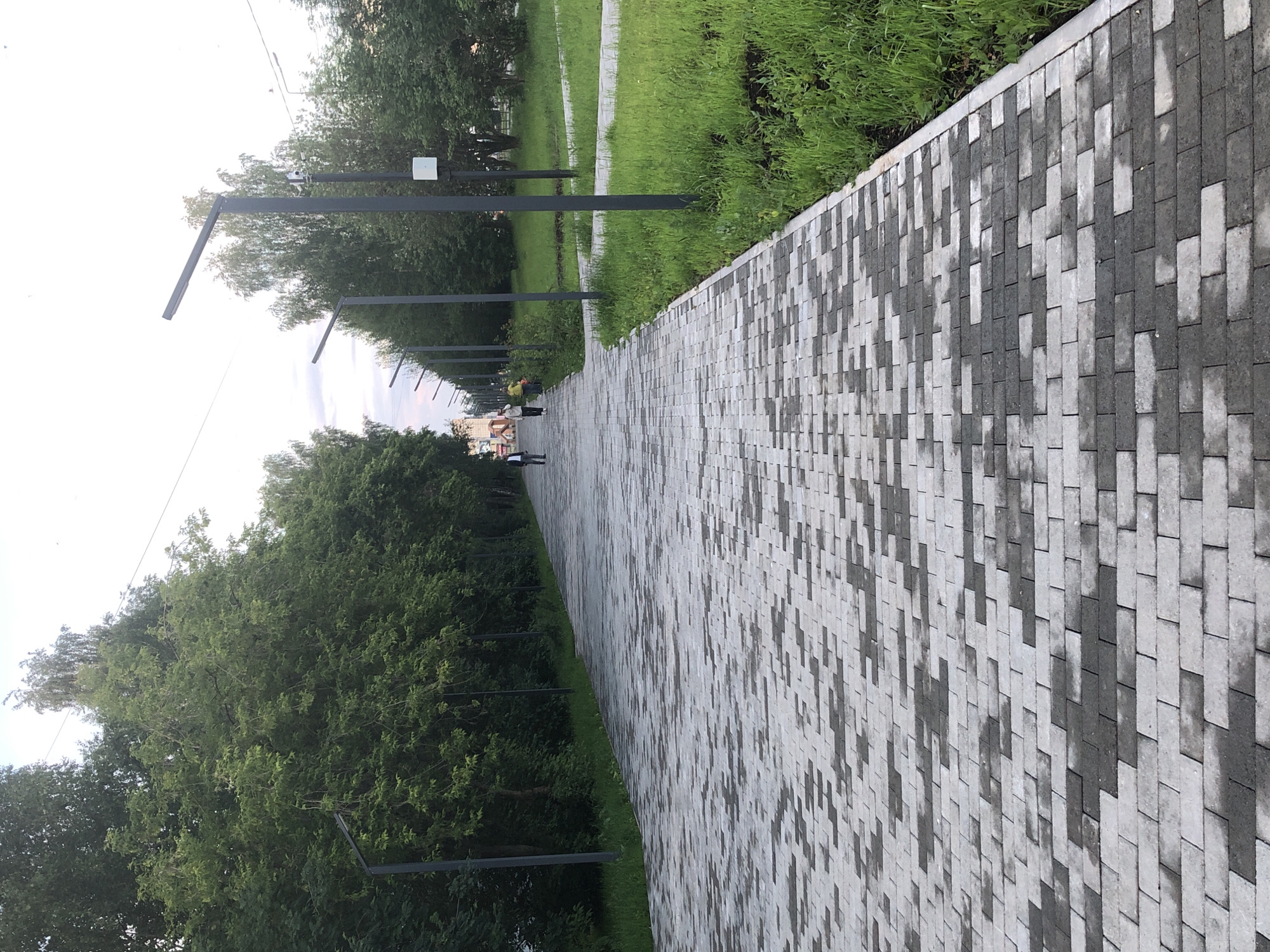 The image shows a paved walkway in a park in Solikamsk, Russia. The walkway is made of rectangular paving stones in varying shades of gray and white, creating a patterned effect. On the left side of the walkway, there is a dense line of green trees. On the right side, there is a well-maintained grassy area, with more trees visible in the background. Several modern-looking streetlights with thin poles and rectangular lamps are spaced along the walkway. In the distance, two people are walking on the path, one in a dark suit and the other in lighter clothing, with a third person slightly further ahead in yellow. The sky is overcast with some lighter areas. The overall scene depicts a well-kept public space.
