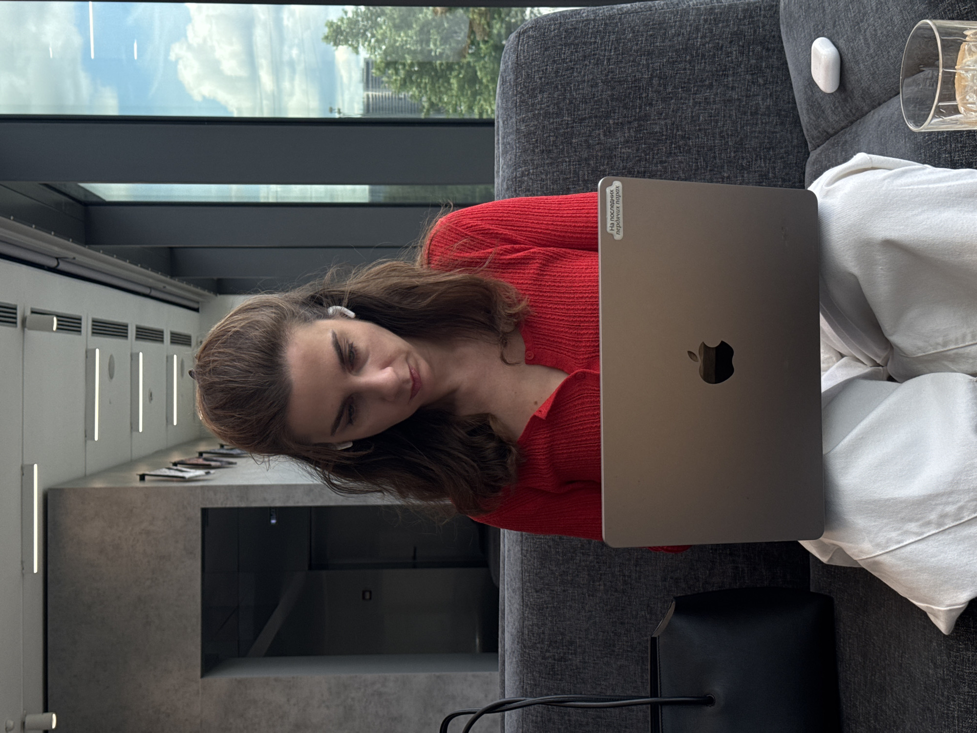 A woman is sitting on a gray sofa in what appears to be an office or lounge setting. She is wearing a red sweater and white pants and has an Apple Macbook laptop in front of her. The laptop has a sticker written in Russian on the upper left corner of the screen. She also appears to have an earbud in her left ear.
Next to her on the sofa is a small white stone and a glass. A black bag is visible next to her laptop. Behind her is a large window with a view of a building and a cloudy sky. The interior walls have a gray concrete finish with vertical light fixtures.
Based on the sticker on the laptop and the architectural style visible through the window, this scene is likely in Moscow, Russia.