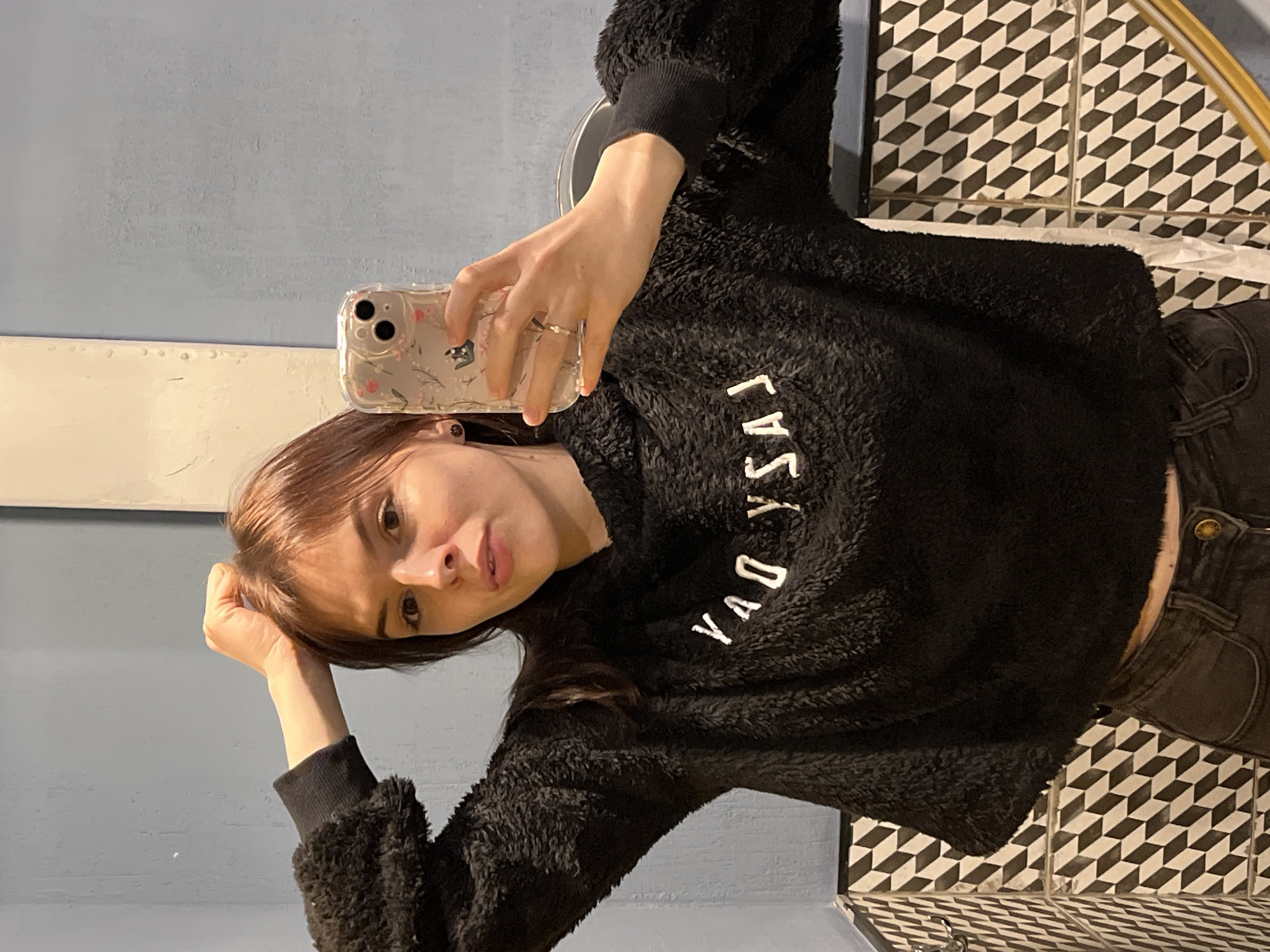 A young woman is taking a selfie in a bathroom. The bathroom is decorated with black and white geometric tiles. The woman is wearing a black fuzzy sweater. Her hair is brown with bangs. The location is Shushary, Russia.