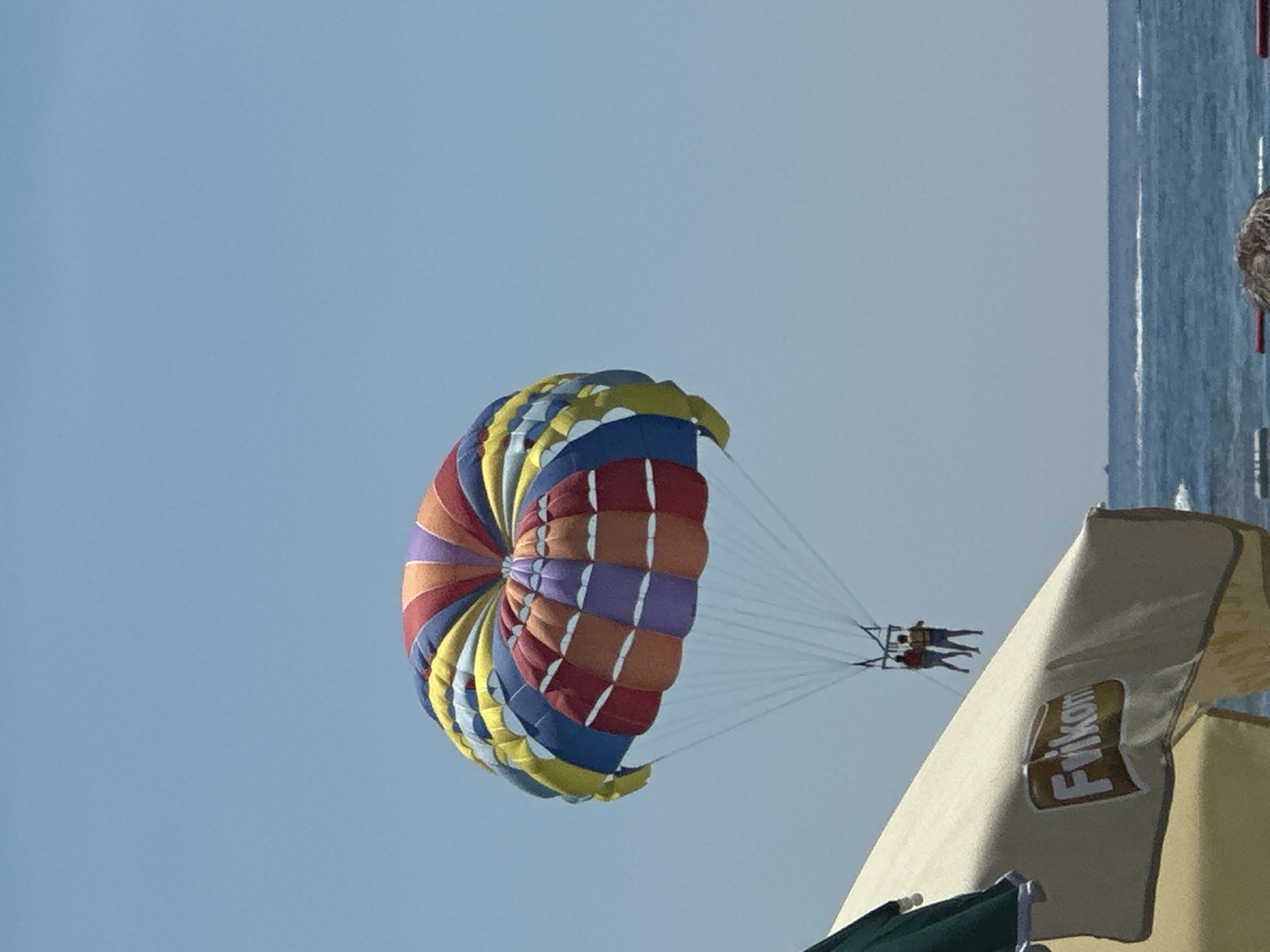 In Boreti, Montenegro, on a clear day, two individuals are parasailing over the Adriatic Sea, suspended from a vibrant, multi-colored parachute. The parachute, adorned with red, orange, yellow, blue, and purple sections, is high in the sky, with thin white ropes connecting it to a harness worn by the parasailers. They are positioned in the mid-ground of the image, with the expanse of the blue sky dominating the upper half. In the foreground, the corner of a large, light-colored beach umbrella is visible, featuring a brown and white logo that reads "Frikam". The sea stretches across the lower portion of the image, with gentle waves and a distant horizon line where the water meets the sky. The overall scene depicts a leisurely summer activity at the coast.