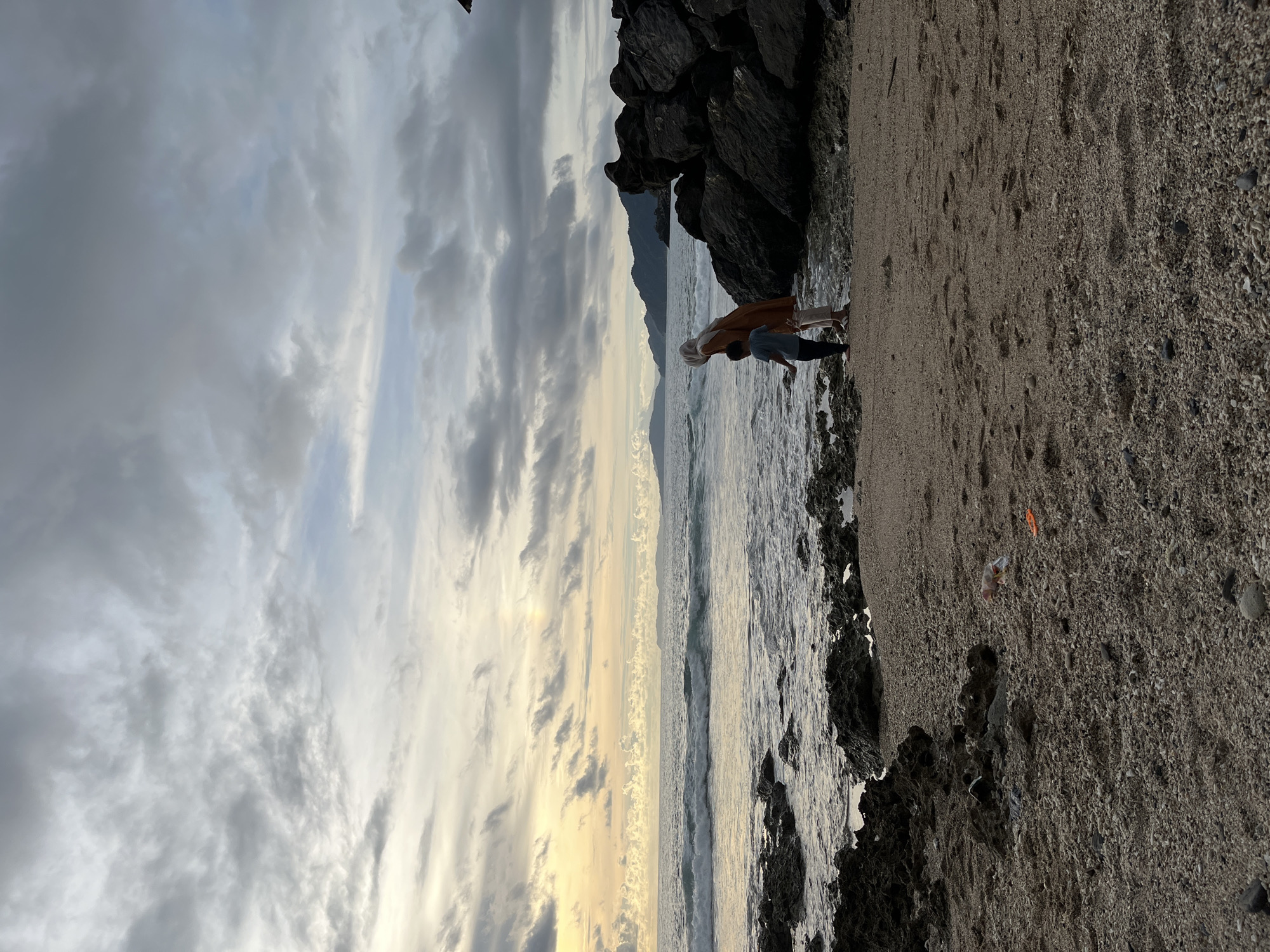 Two people, one wearing a tan garment and head covering, and another in a blue shirt, are standing on a sandy beach at the edge of the water. The scene is a seascape with a rocky shoreline. In the background, mountains are visible behind the ocean. The sky is filled with dramatic clouds, with patches of sunlight breaking through, suggesting either sunrise or sunset. The overall mood appears calm and contemplative. The image is oriented vertically.