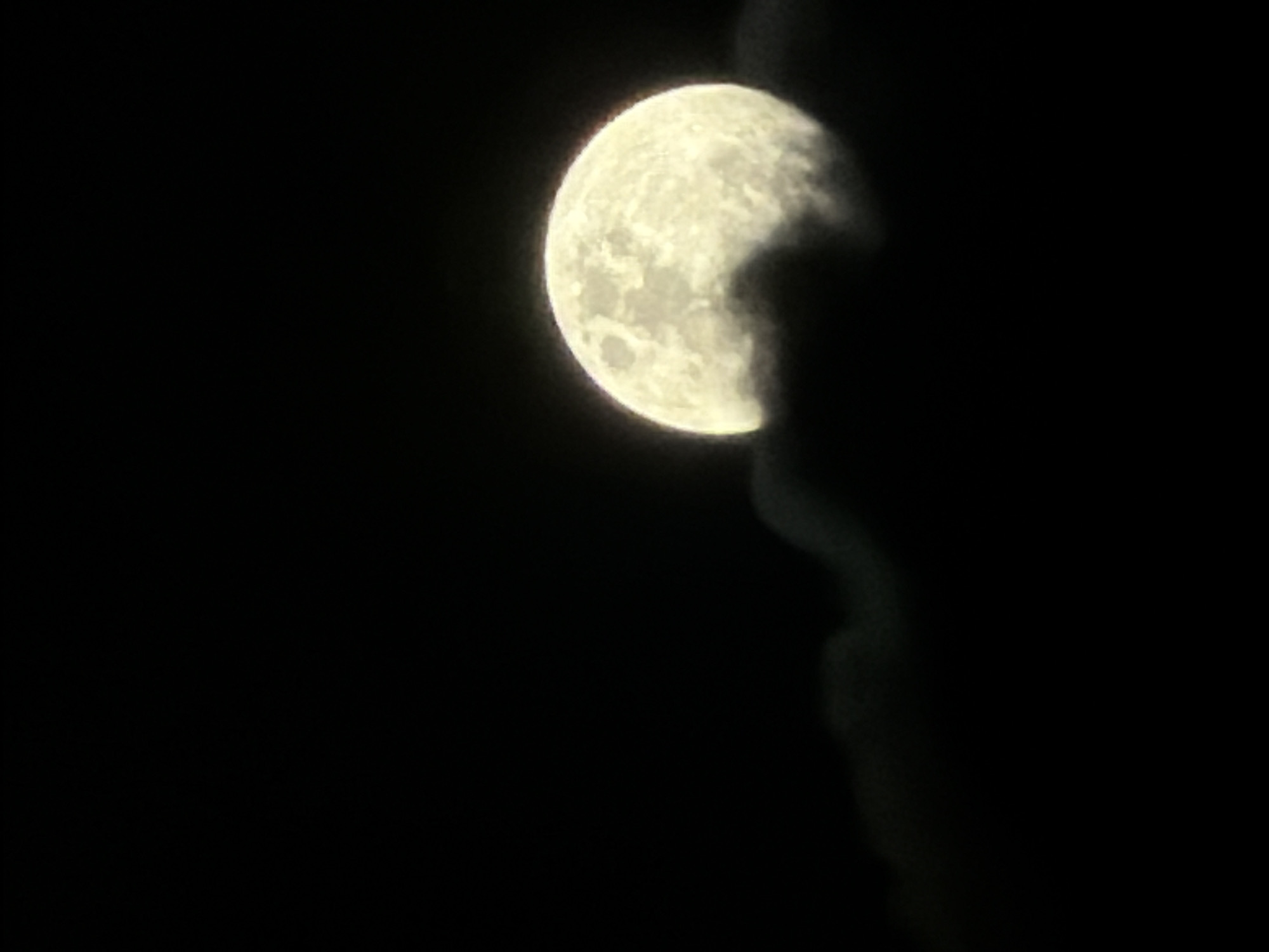 The image displays a close-up view of the Moon in a dark night sky, partially obscured by dark, wispy shapes that appear to be clouds or atmospheric haze. The Moon, appearing yellowish-white and brightly illuminated, occupies the upper-middle section of the frame, with approximately three-quarters of its surface visible. Its craters and surface textures are discernible, showing variations in brightness across its disc. The dark, amorphous forms intrude from the right side of the image, covering a portion of the Moon and extending downwards into the blackness of the sky. The background is entirely black, indicating the absence of other light sources or celestial bodies. The overall impression is one of a celestial observation under veiled conditions at night. No people or visible text are present. This scene was captured in North Kuta, Indonesia.