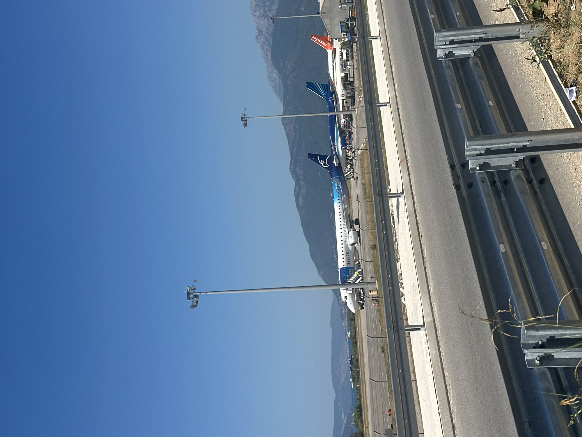 The image shows an airport tarmac with several airplanes parked. The sky is clear and blue. In the foreground, there is a road and a metal guardrail. In the mid-ground, there is a tarmac with several airplanes, including a white and blue airplane with "Air Montenegro" written on it, a blue airplane with a bird logo, and an orange airplane with "easyJet" written on it. There are also airport lights and some people standing near the airplanes. In the background, there are mountains and some buildings. The scene appears to be a sunny day at an airport. The location is Tivat Airport in Mrčevac, Montenegro.