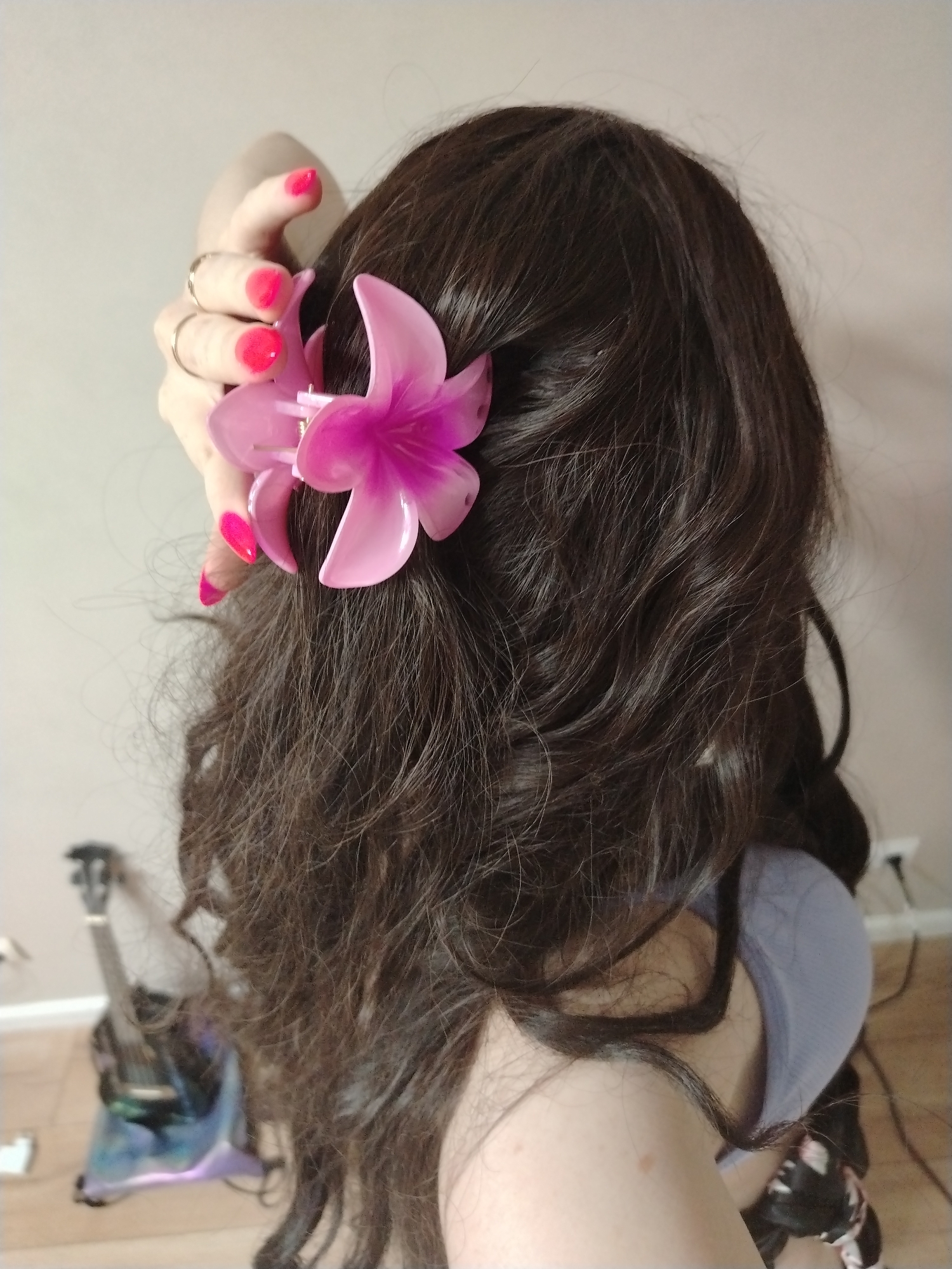 A person with long, wavy, dark brown hair has a pink flower clip in their hair, near their right ear. Their hand, with pink nail polish and rings on the fingers, is holding the clip. They appear to be wearing a light purple top. In the background, out of focus, is a guitar stand and a guitar. The image was taken indoors against a light-colored wall and a hardwood floor. There is no information to ascertain whether this image was taken in Izhevsk, Russia.