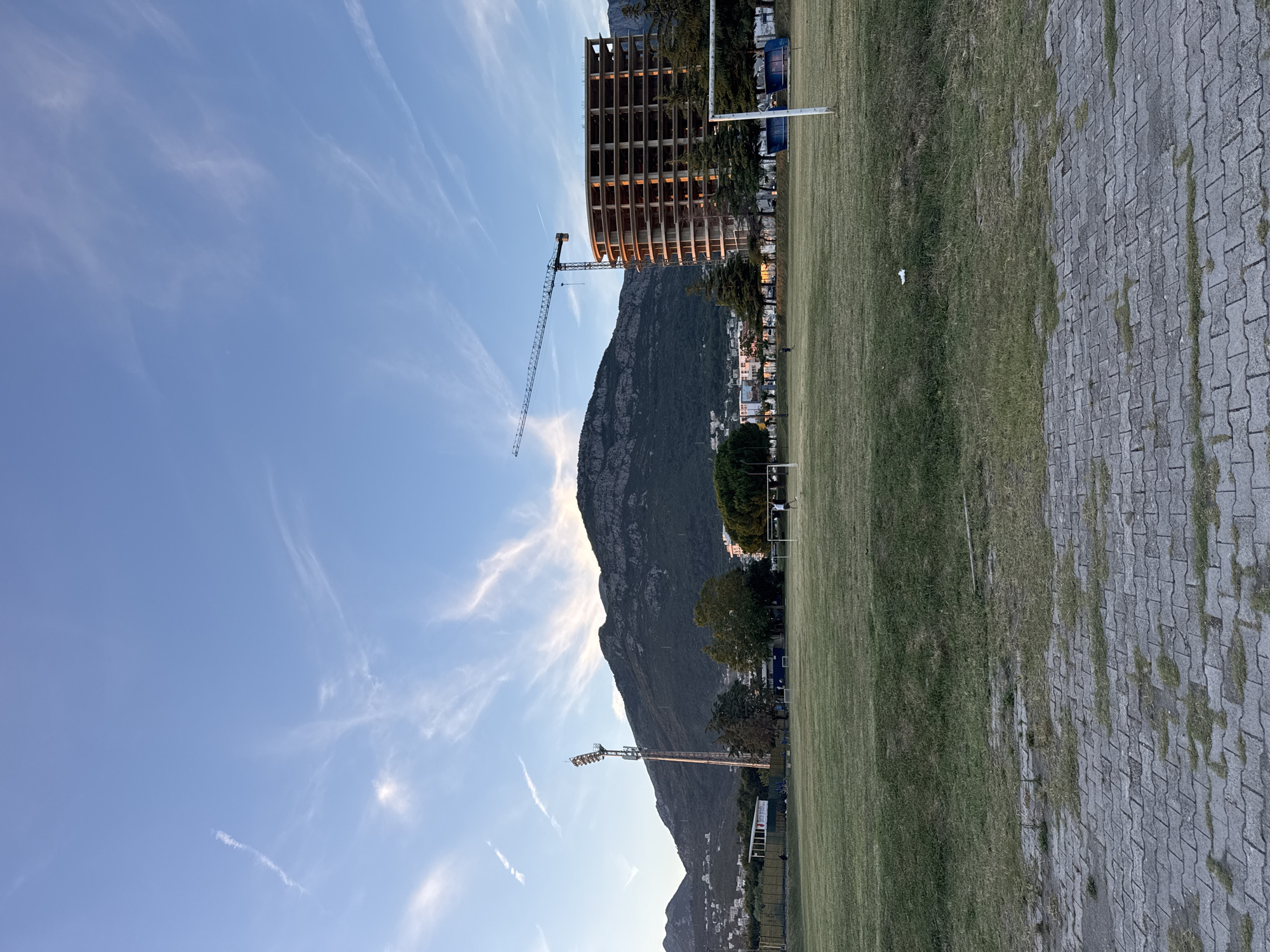 This image captures a wide-angle view of a football pitch and its surroundings in Bar, Montenegro, during late afternoon or early evening. The foreground features a paved area of gray interlocking bricks with sparse grass growing between the cracks, transitioning into a large, green but somewhat dry grassy field that constitutes a football pitch. Two white goalposts are visible, one partially on the far right and another less defined in the center-right midground. A solitary individual, a human male, appears near the right goalpost, facing away from the viewer. Another smaller figure is visible further back on the field towards the center.
In the midground on the left, a tall stadium light tower equipped with multiple floodlights stands prominently, illuminated by the low sun from the right. Behind the light tower and extending across the left side of the frame, forested mountains with scattered white residential buildings on their lower slopes rise against the sky. The dominant feature in the background is a massive, dark, rugged mountain range, likely the Rumija mountain range, with visible rock faces and patches of vegetation, framing the scene.
On the right side of the midground, a large, multi-story, curved or oval-shaped building is under construction, its concrete frame exposed and partially lit by the sun. A tall construction crane with a horizontal jib extends from behind this building, its counterweight block visible. Several coniferous trees are planted in a row in front of the construction site, partially obscuring the lower levels.
The sky is a clear blue with wispy, elongated cirrus clouds stretching across it, indicating fair weather with subtle atmospheric movement. The light suggests either a sunset or sunrise, with warm tones on the clouds and structures. No direct interactions between individuals are discernible.