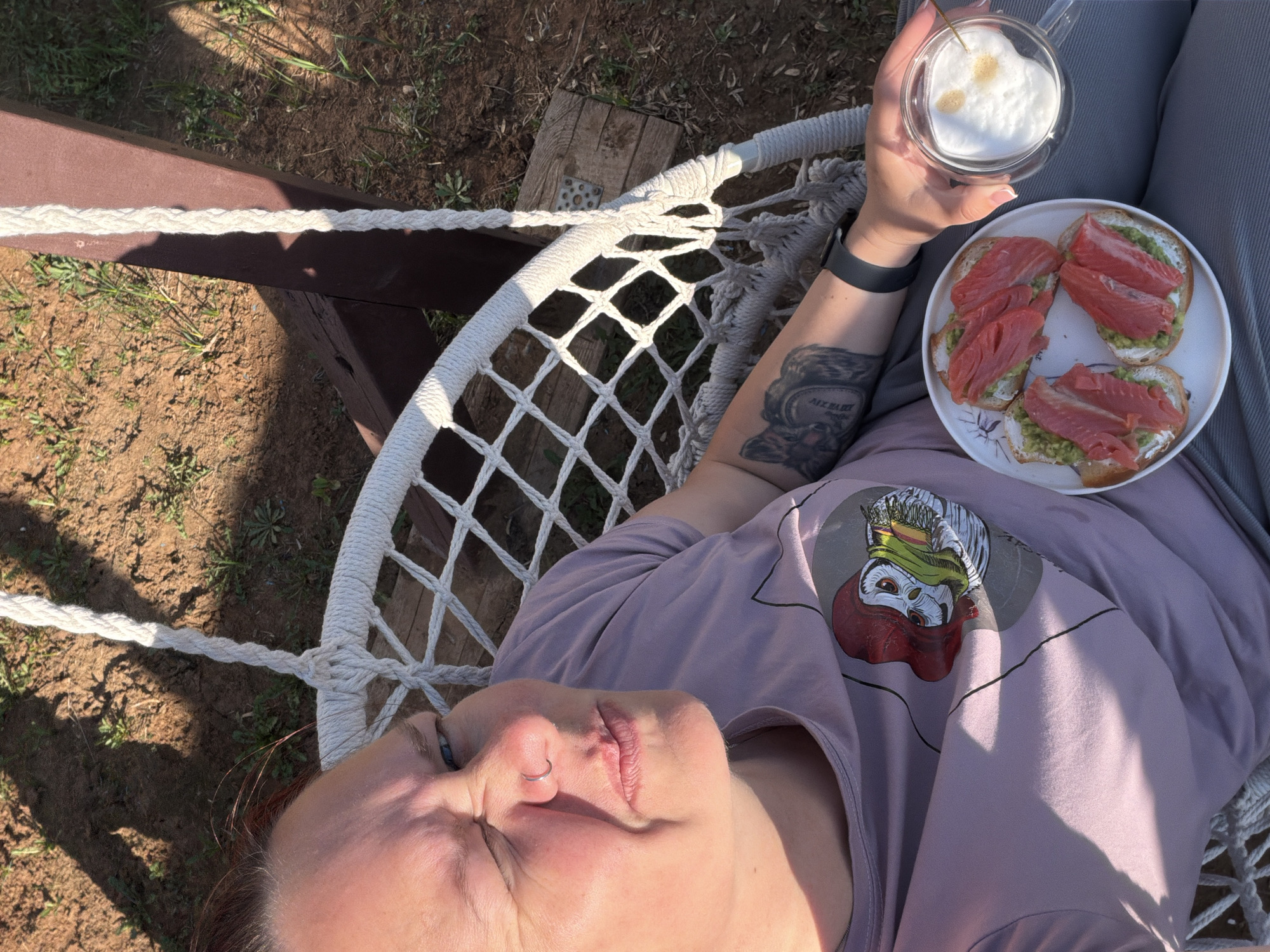 A woman is reclining in a white rope hammock outdoors, likely in a backyard or garden, as suggested by the dirt and vegetation in the background. She's wearing a lavender t-shirt with a graphic design and a black watch on her left wrist. She is holding a clear glass mug with a frothy beverage in her right hand and a white plate with three pieces of toast topped with what appears to be avocado or another green spread, and salmon or smoked salmon. She has a nose piercing. Based on the available information, a specific location in the city of Chaikovsky, Russia is not determinable from the image.