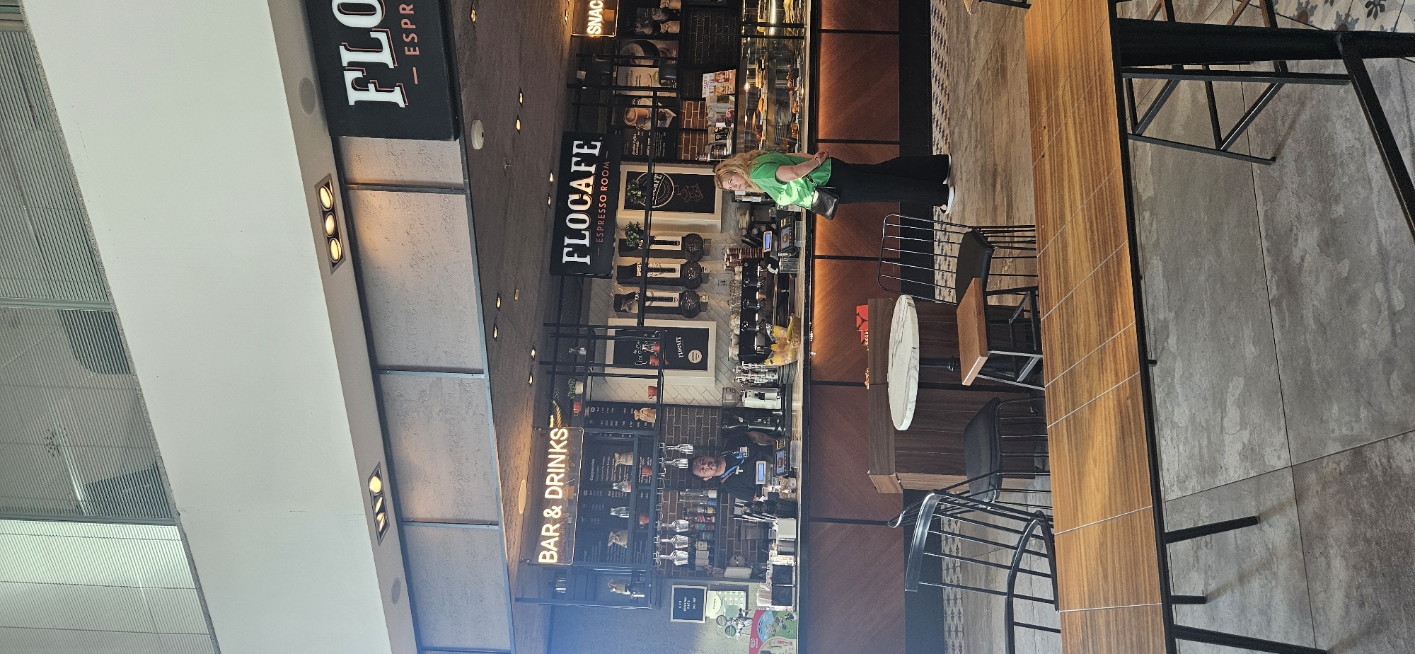 The image depicts the interior of a modern cafe with a counter service area. The counter is made of wood with a dark brown facade and is illuminated by warm-toned LED lighting underneath. Behind the counter, shelves are stocked with various items, including glassware, mugs, bags of coffee, and decorative plants. A menu board is prominently displayed, listing coffee and drink options. On the right side of the counter, a glass display case showcases pastries and other food items.
A male employee, wearing a dark shirt and a lanyard with an ID badge, stands behind the counter, appearing to be serving customers or preparing orders. A female customer, dressed in a bright green top and black pants, stands in front of the display case, looking to her left.
The cafe has a contemporary design, with metal chairs and tables in the foreground. The floor is a mix of concrete tiles and decorative patterned tiles. Above the counter, illuminated signage reads "BAR & DRINKS" and "FLOCAFE - ESPRESSO ROOM". Another sign, partially visible on the far right, says "SNACKS". The ceiling features industrial-style lighting fixtures. The overall atmosphere suggests a busy and stylish cafe environment. This cafe is located in Budapest, Hungary.