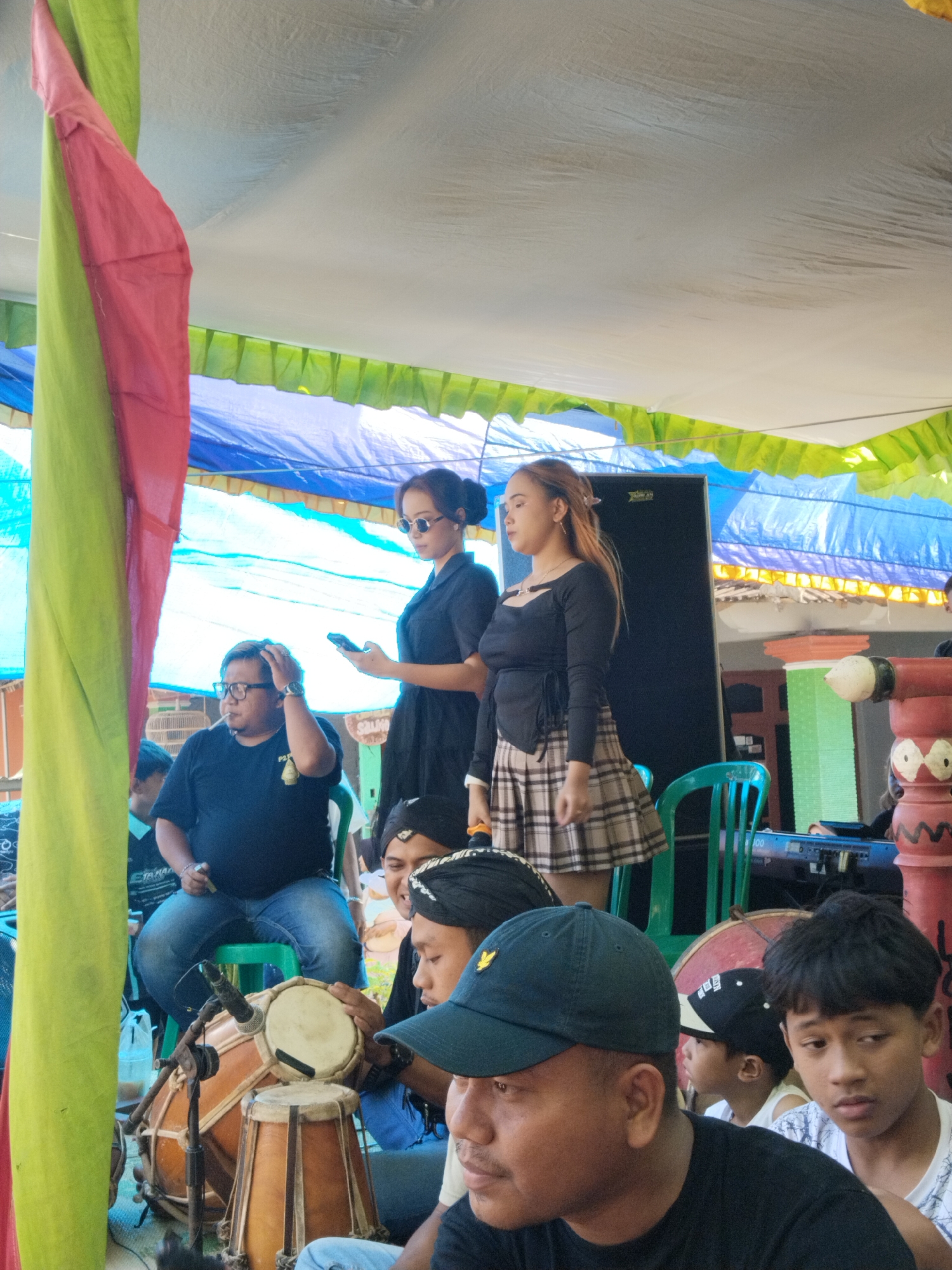 The image shows a group of people gathered under a tent-like structure in Mojodoyong, Indonesia. The tent is decorated with red, green, and blue fabric. A man in a black shirt and jeans sits on a green stool, holding his head. Next to him, a woman in sunglasses and a black dress holds a mobile phone. Another woman with long hair, wearing a black top and a plaid skirt, stands beside her. In the foreground, several men are seated, some wearing hats. One man is playing a pair of drums. A keyboard is visible in the background next to a decorative pole. The overall impression is that of a casual gathering or performance.