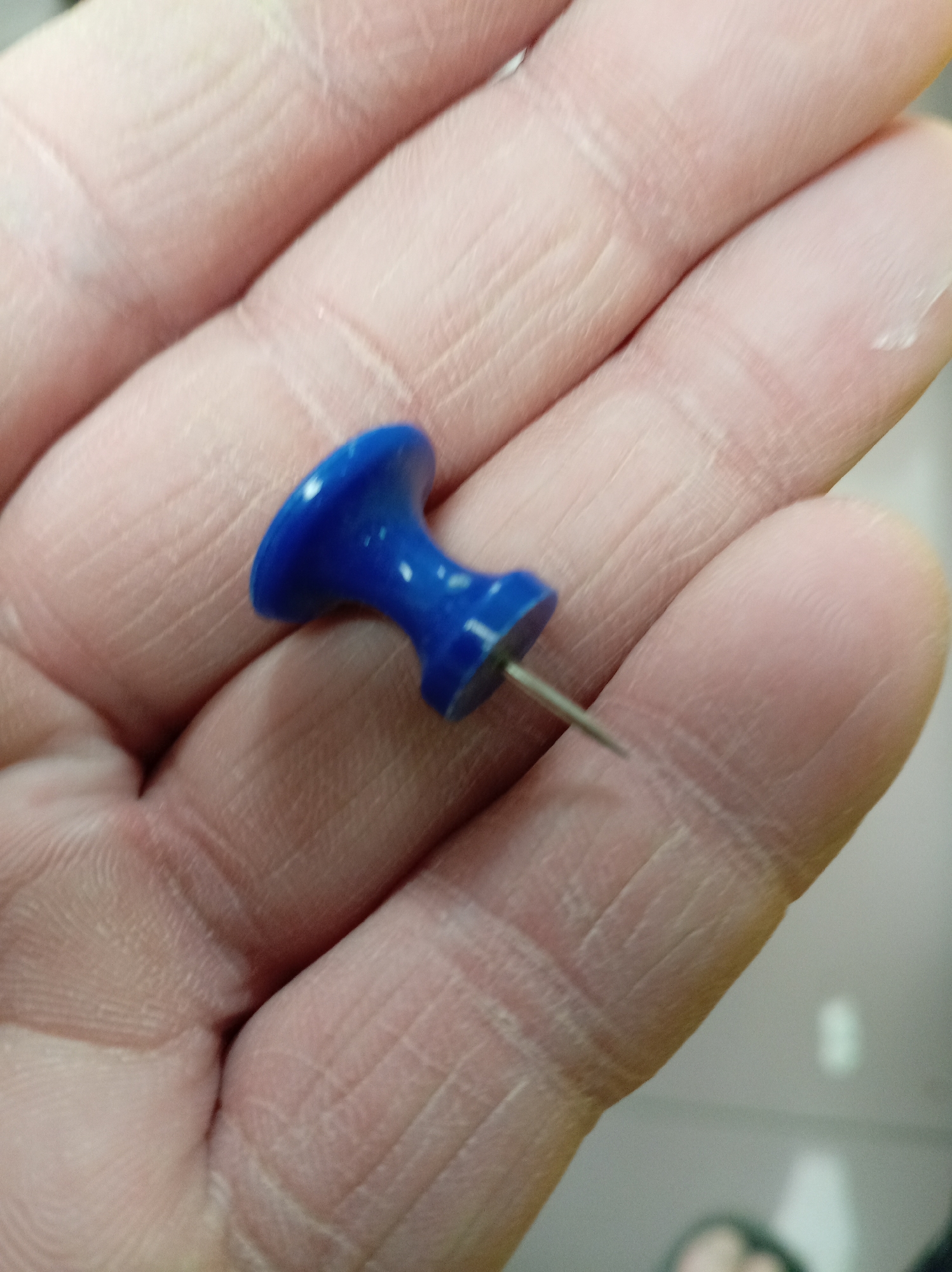 A close-up image shows a blue pushpin being held in a person's palm. The pushpin's point is visible. The person's hand appears weathered. The background is blurry and indistinct. Location is unknown.