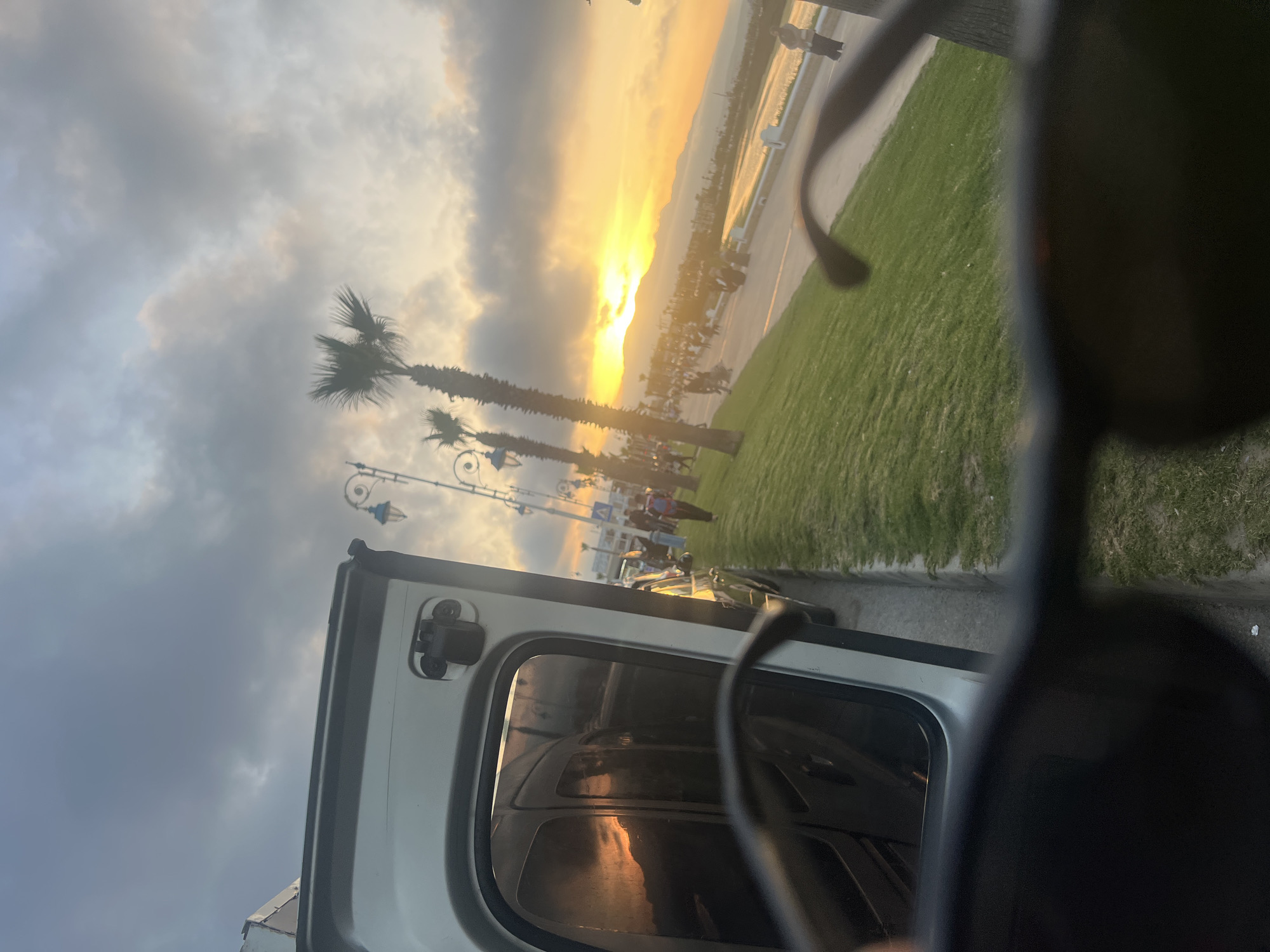 The image, taken in Martil, Morocco, shows a sunset scene from what appears to be inside a vehicle, partially obscured by a pair of sunglasses. Palm trees stand along a grassy area adjacent to a road or path. The sunset illuminates the clouds and reflects in the vehicle's window. People are walking on the road or path. Vintage street lights are visible among the trees. The vehicle door is open.